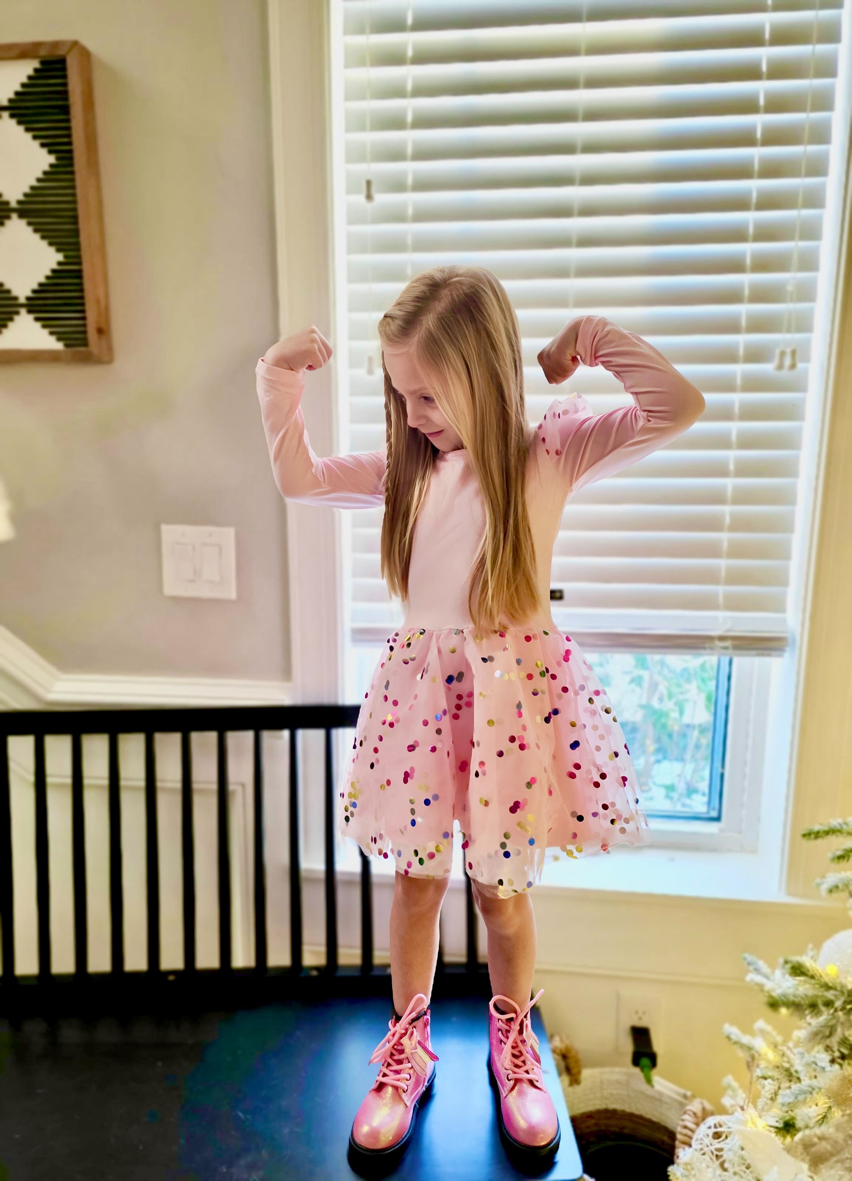 Girl in pink dress and boots flexing arms near window.