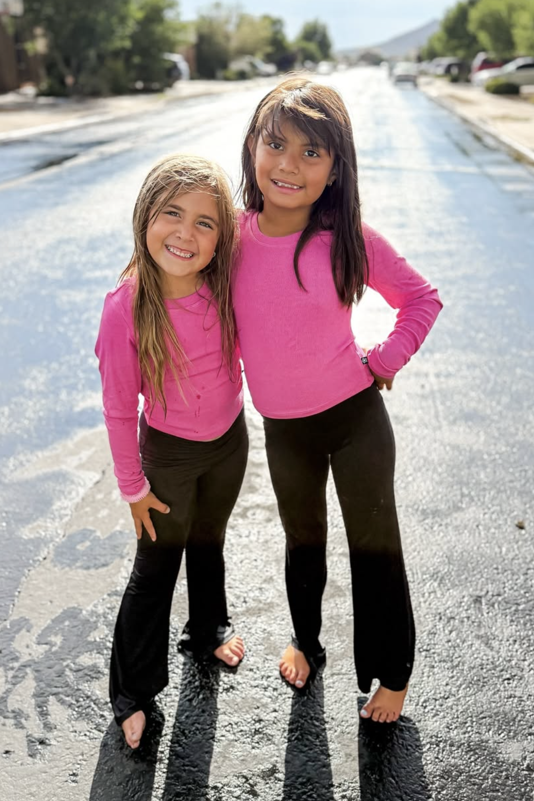 Two children in pink shirts and black pants stand barefoot on a wet street.