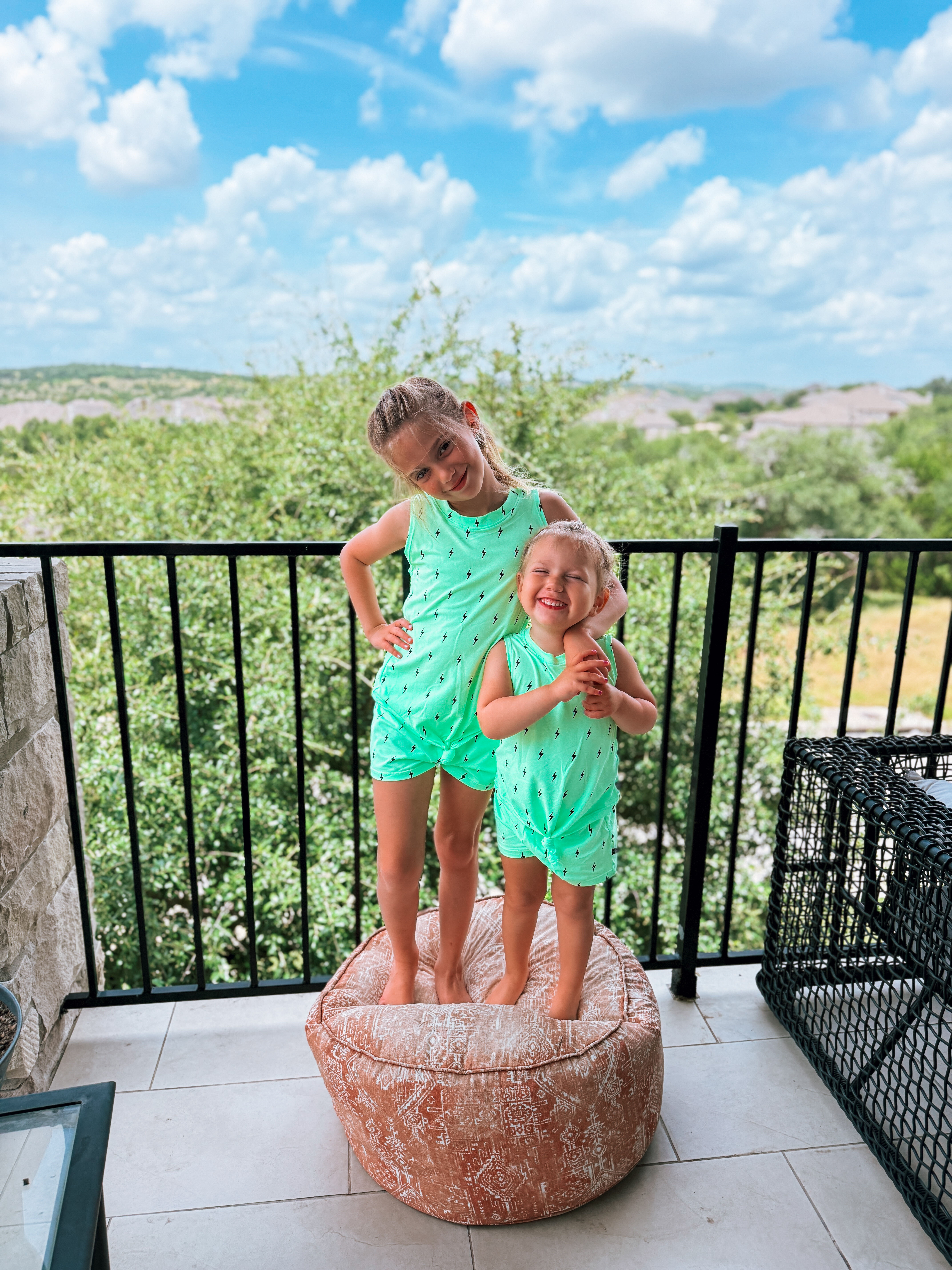 Two children in matching outfits stand on a cushion on a balcony, scenic view behind them.