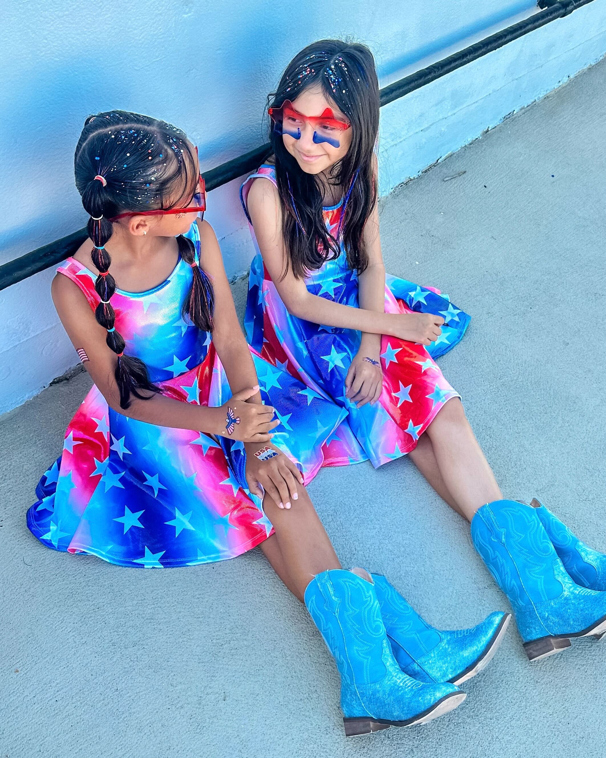 Two girls in star-patterned dresses and blue boots sitting on pavement.
