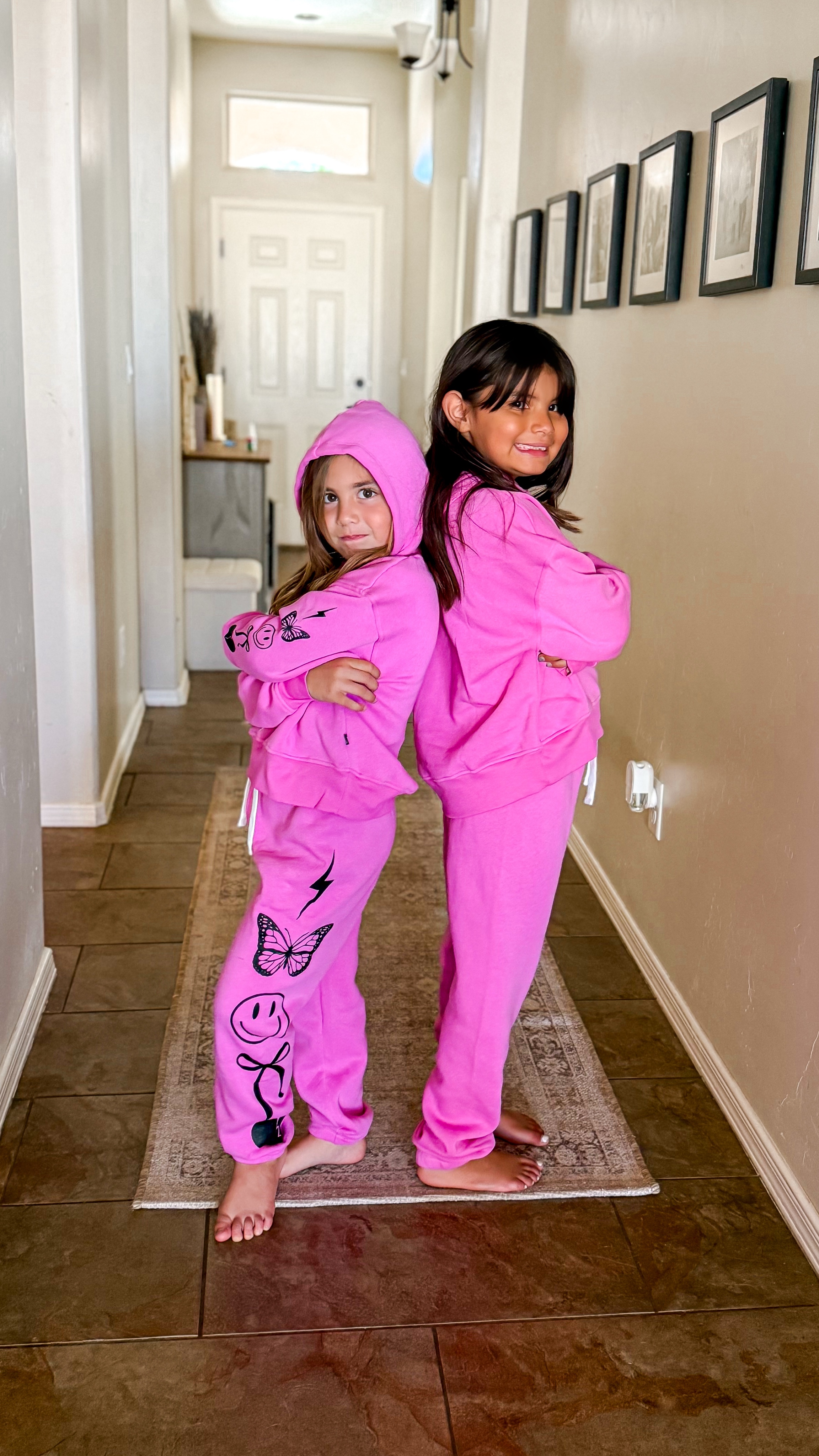 Two children in pink hoodies pose back-to-back in a hallway.