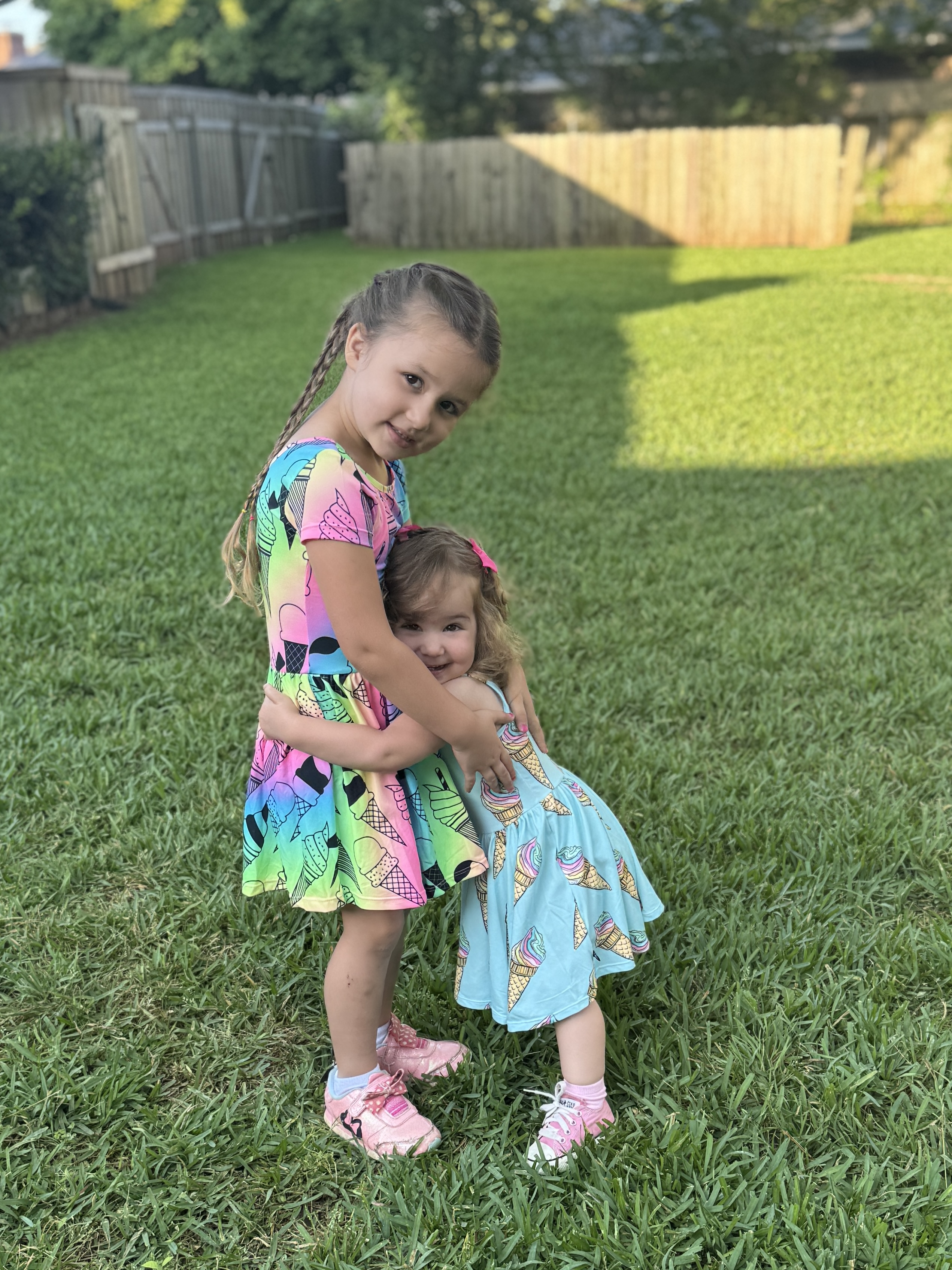 Two young girls in colorful dresses hugging on a grassy lawn.