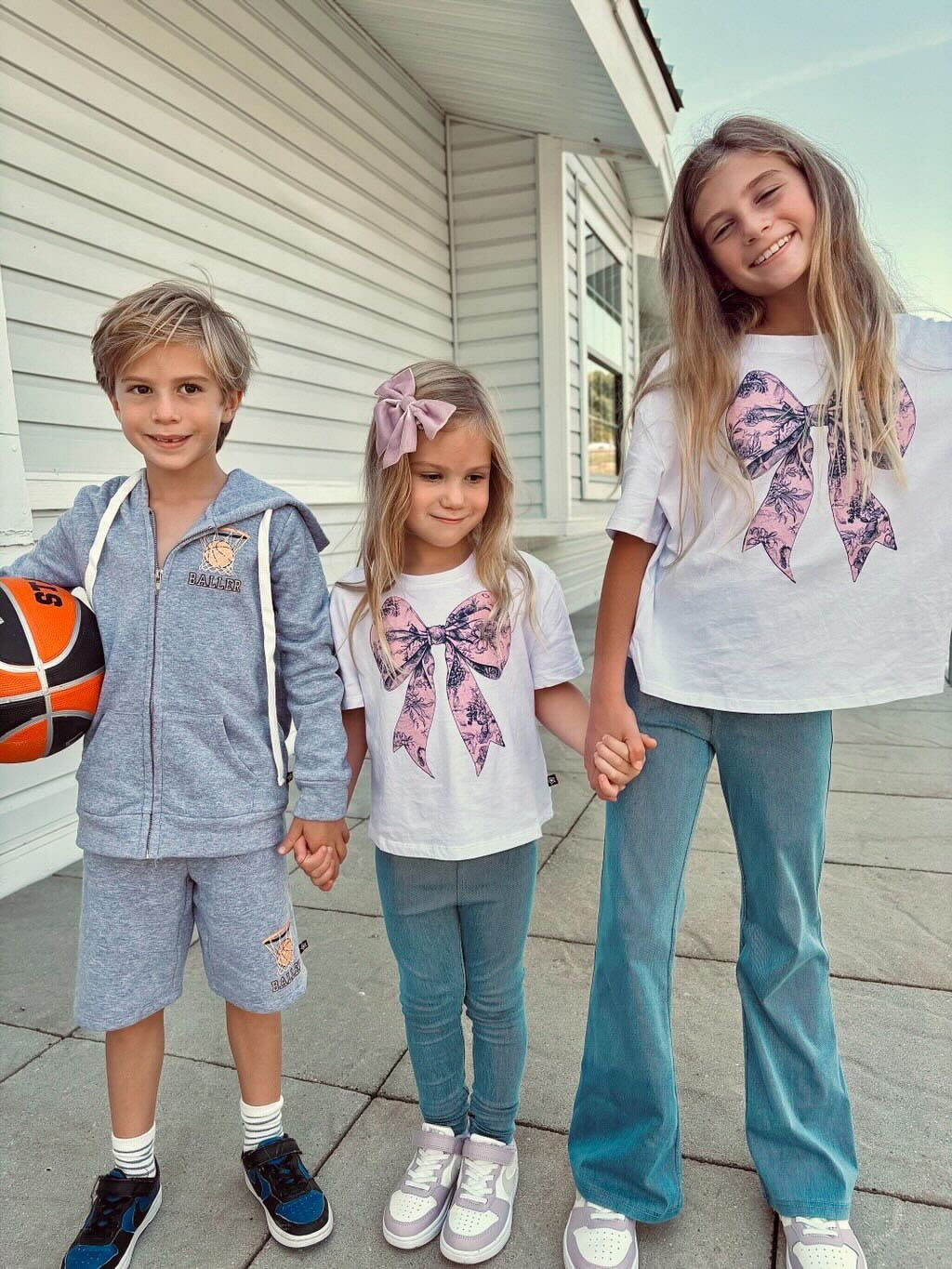 Three children standing outdoors, holding hands, smiling, one holds a basketball.