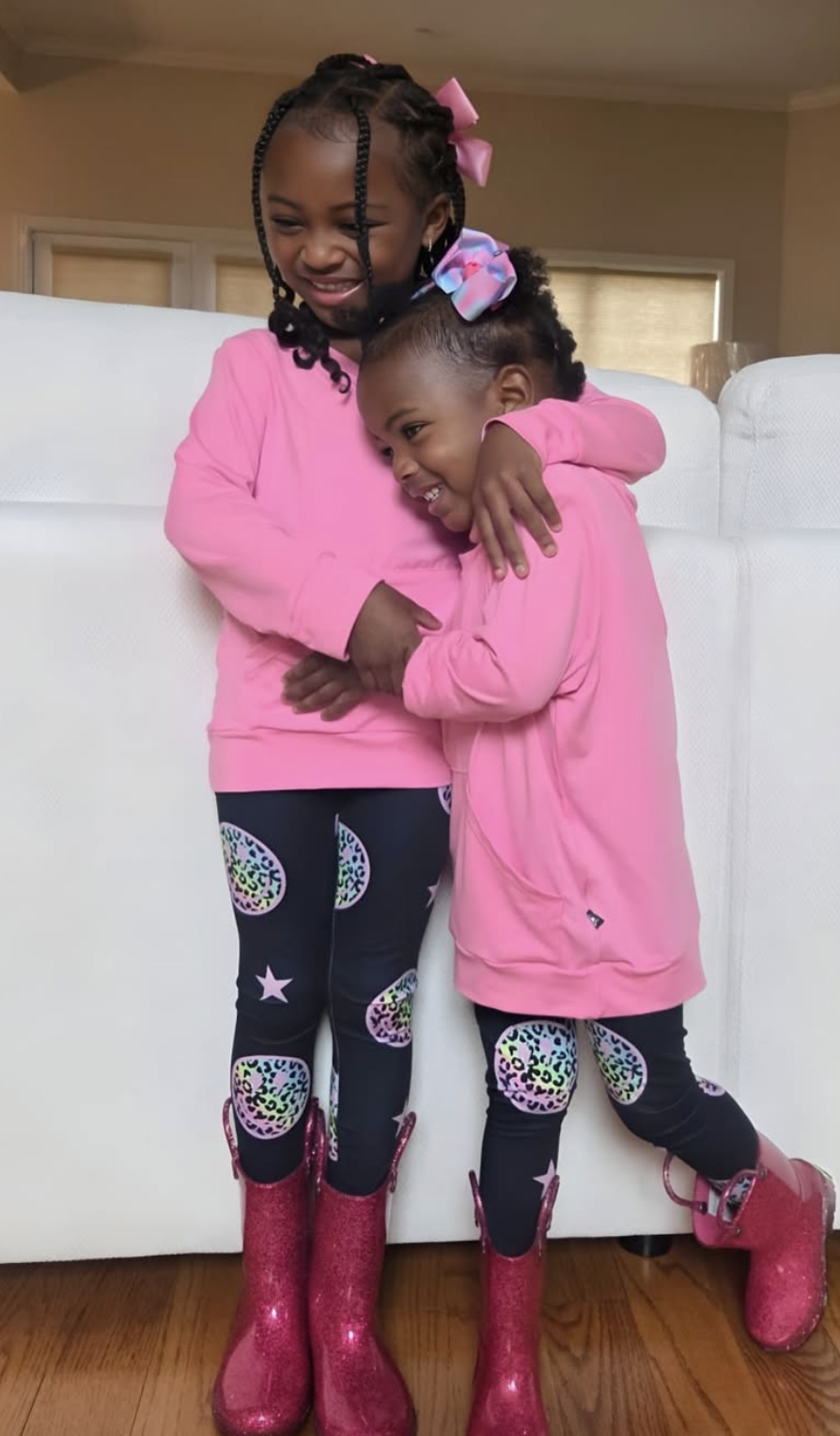 Two children in pink outfits hugging, wearing matching pink boots.