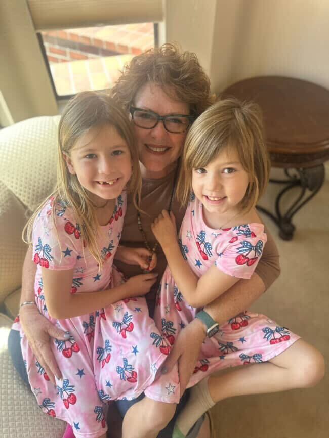 Person sitting with two children in matching pink dresses with patriotic designs indoors.