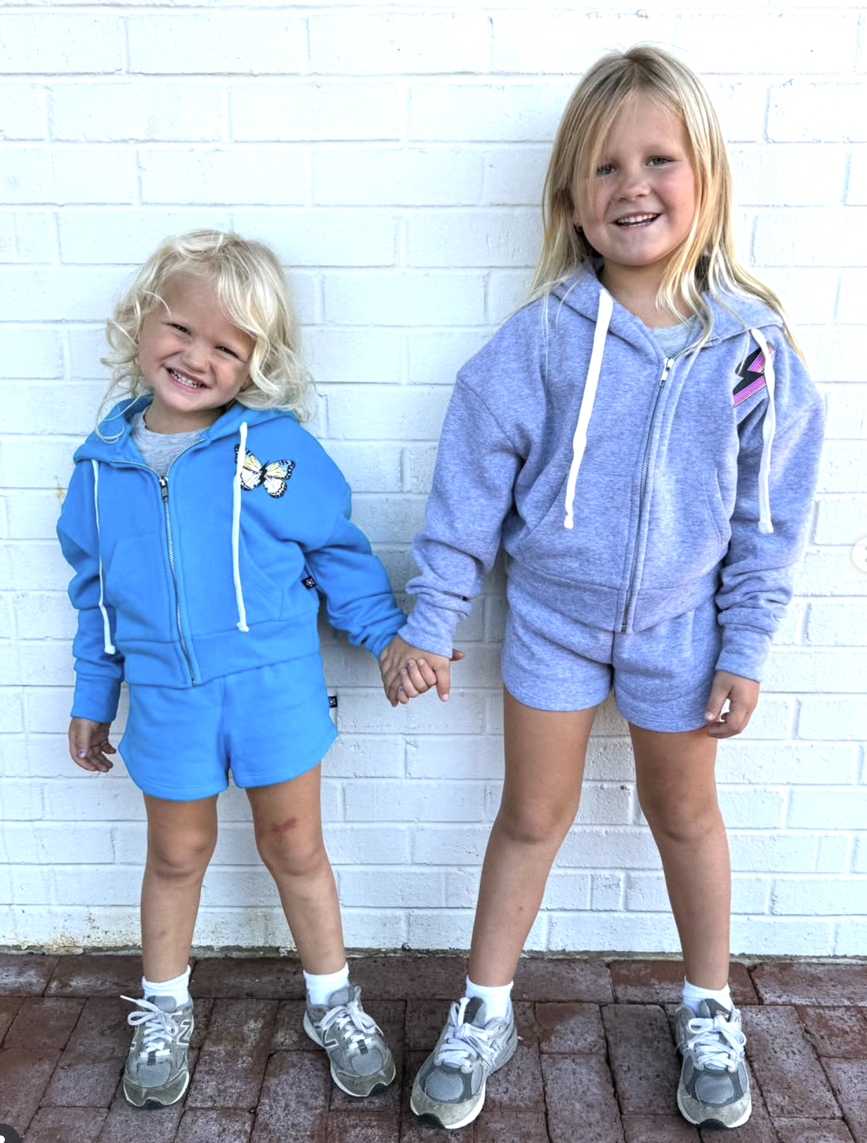 Two children in colorful outfits smiling and holding hands against a white brick wall.