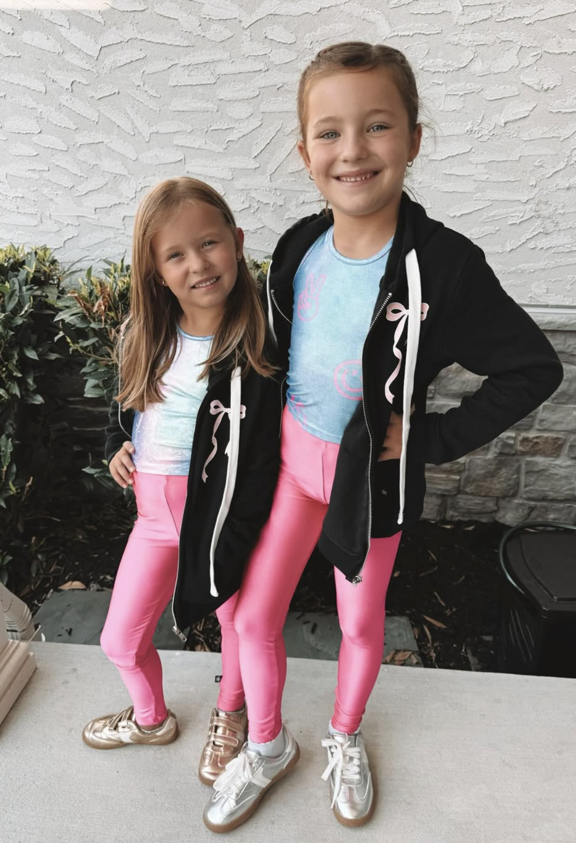 Two children in matching outfits with pink pants and black hoodies, standing outside.