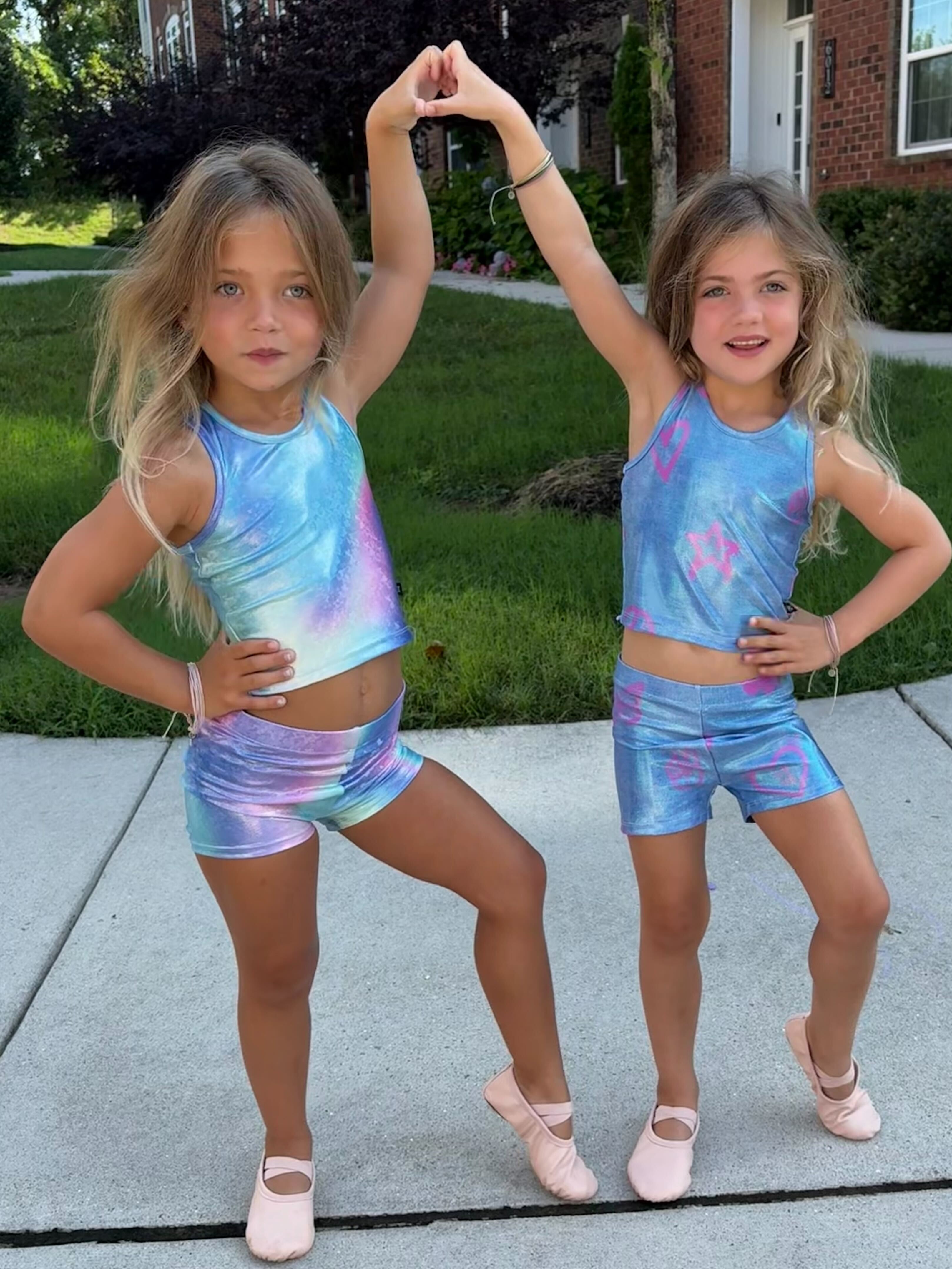 Two young girls in colorful dance outfits pose outside on a sidewalk.