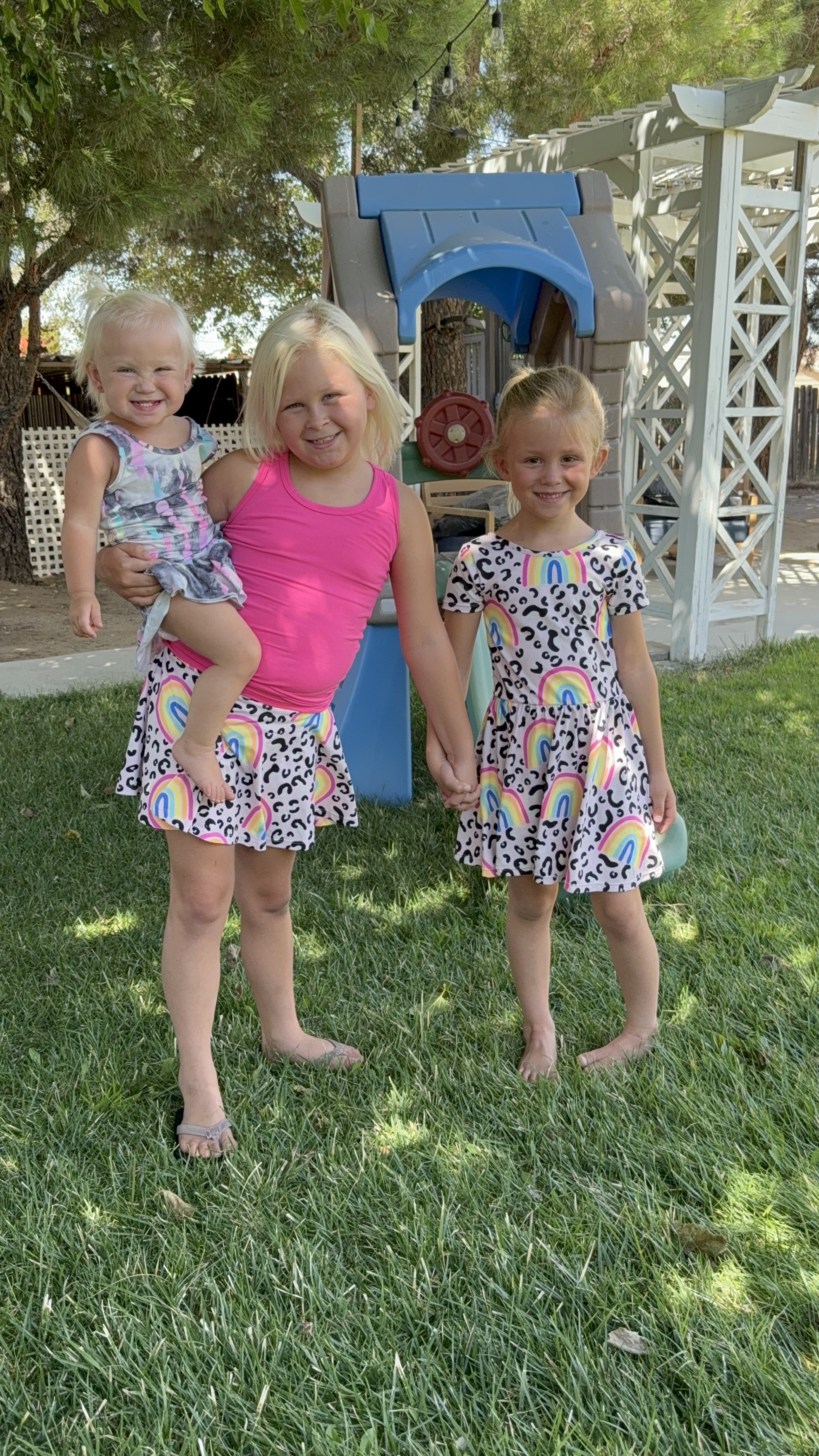 Three children smiling, standing on grass in a backyard with a playhouse in the background.