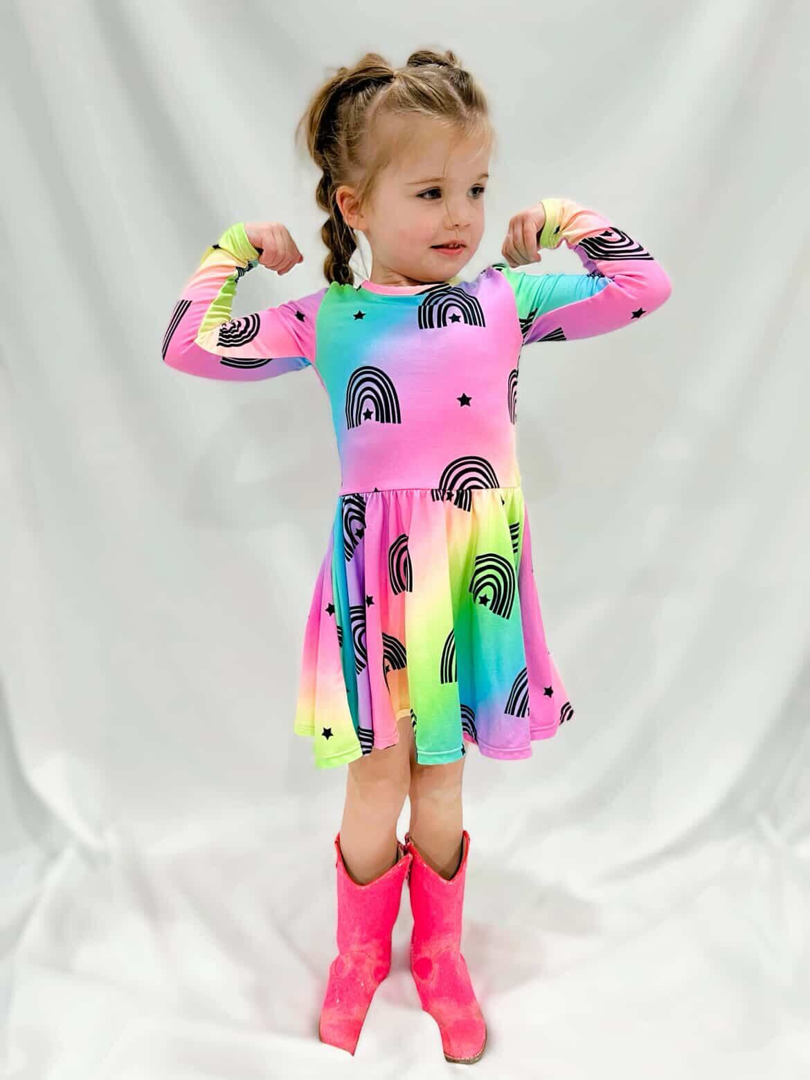 Child in colorful rainbow dress and pink boots, posing with arms flexed.