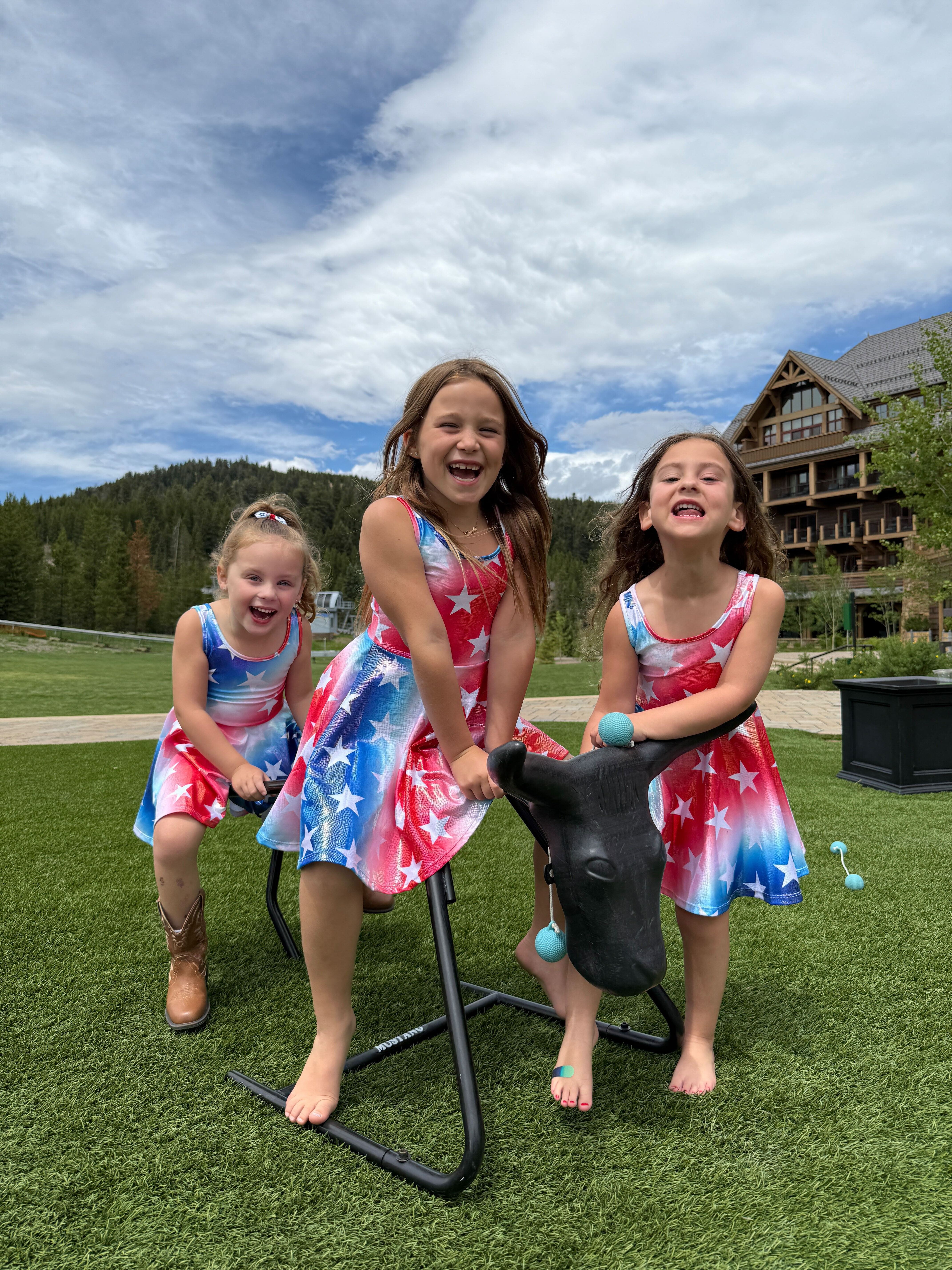 Three girls in matching star-patterned dresses, playing outside near a large wooden building.