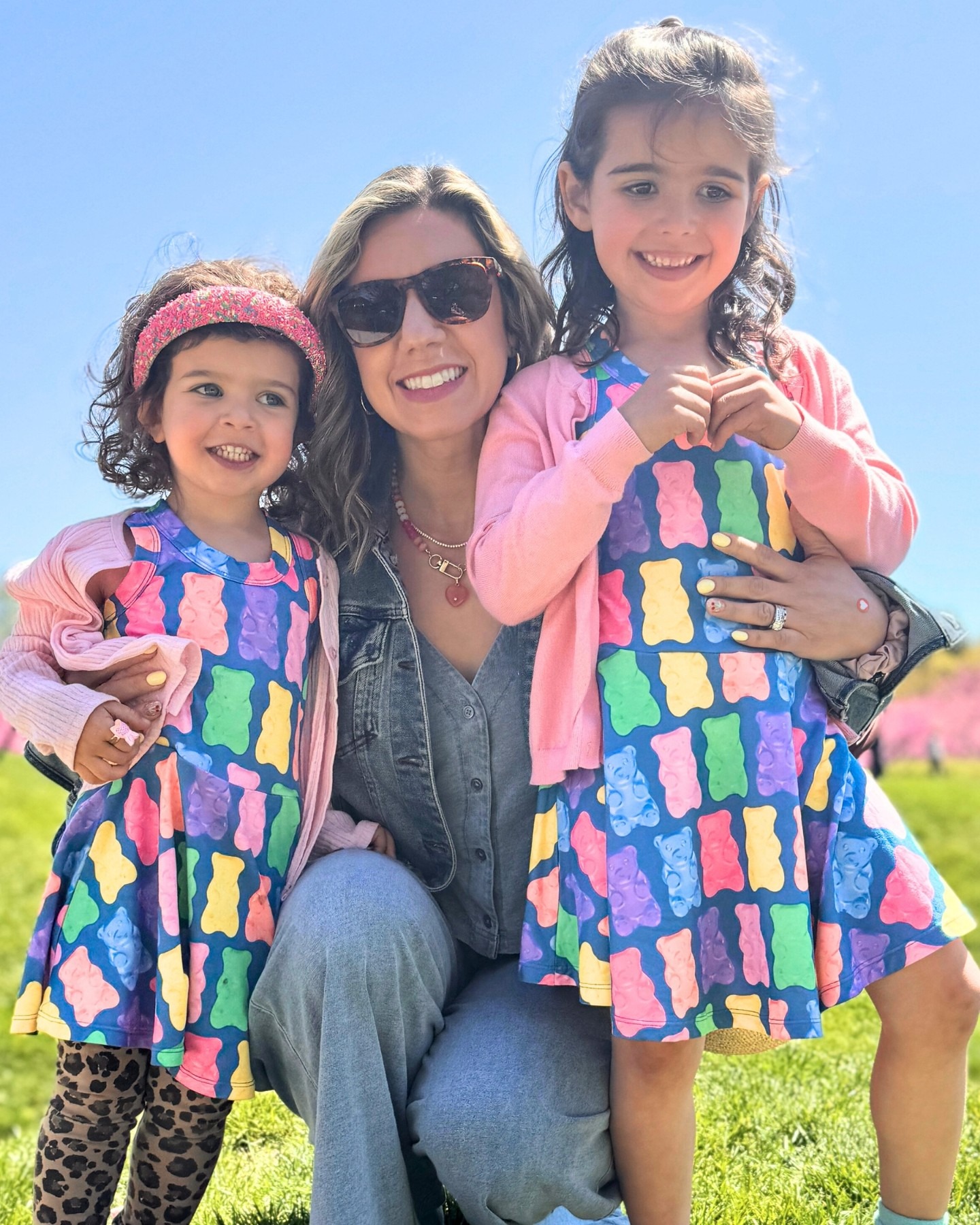 A woman with sunglasses and two children in colorful dresses smiling outdoors.
