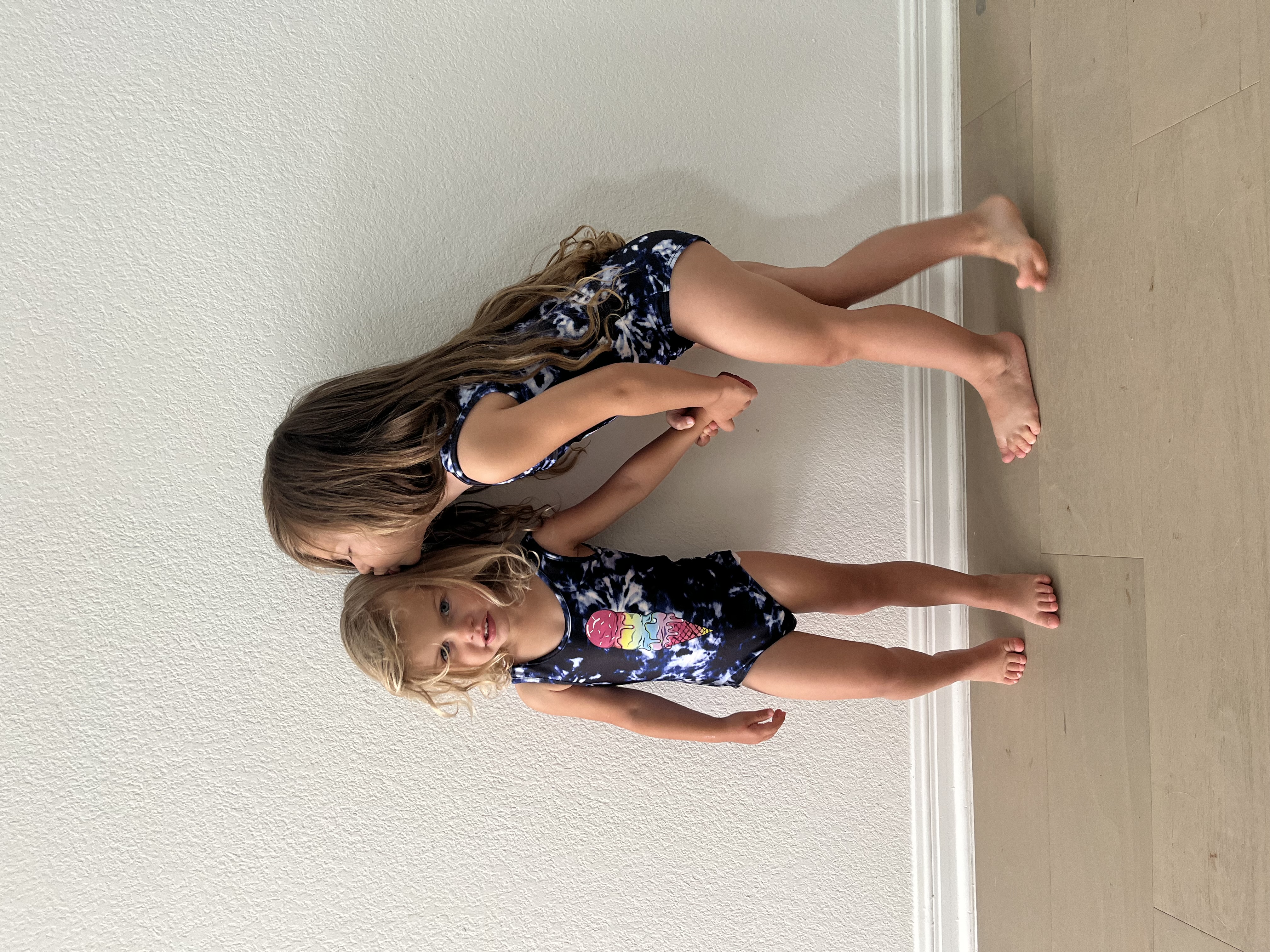 Two children in matching swimsuits, one kissing the other's head, standing against a white wall.