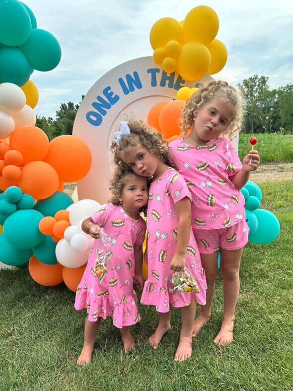 Three children in matching pink outfits standing by colorful balloon decorations outdoors.