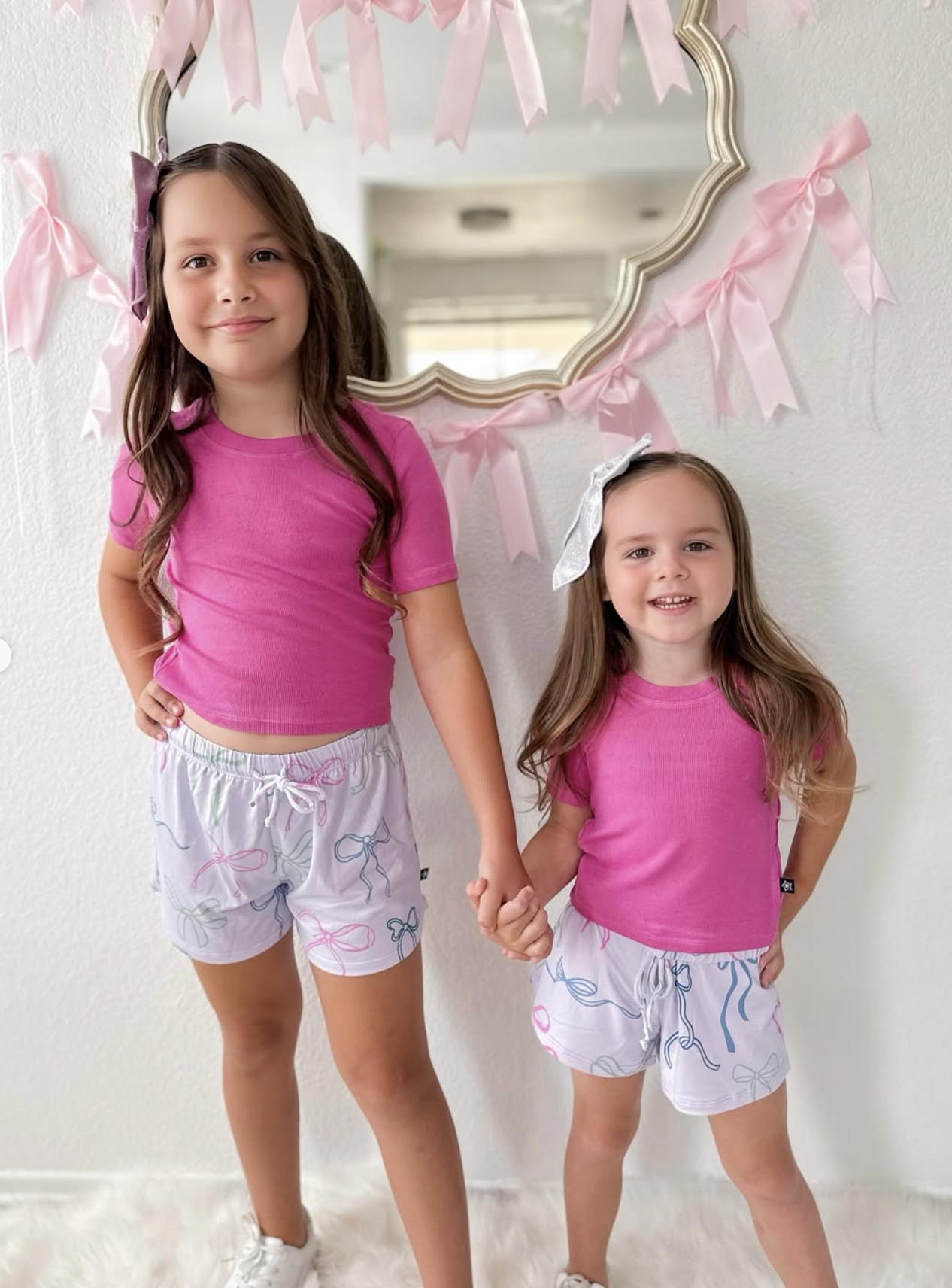 Two children in matching pink shirts and shorts, holding hands, in front of a decorated mirror.