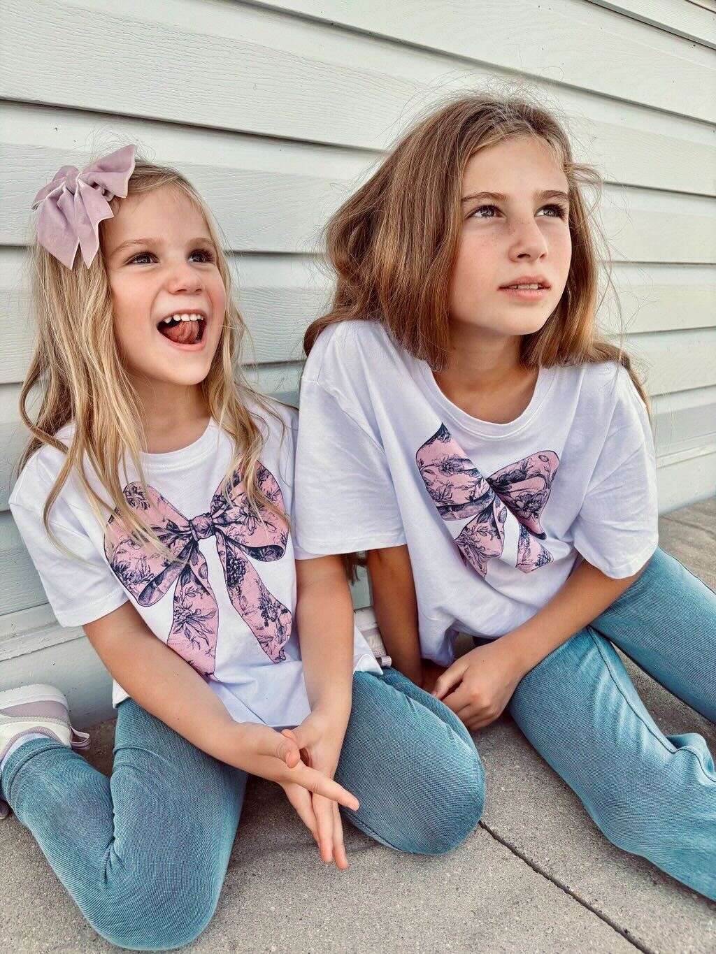 Two children sitting near a wall, wearing matching shirts with bows.