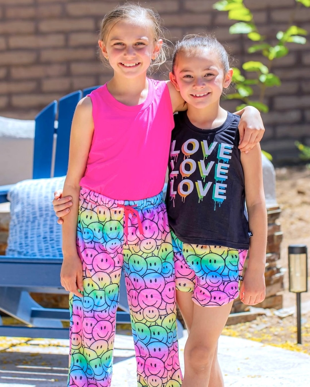 Two children smiling, wearing colorful smiley face pants and a 'LOVE' shirt, outdoors.