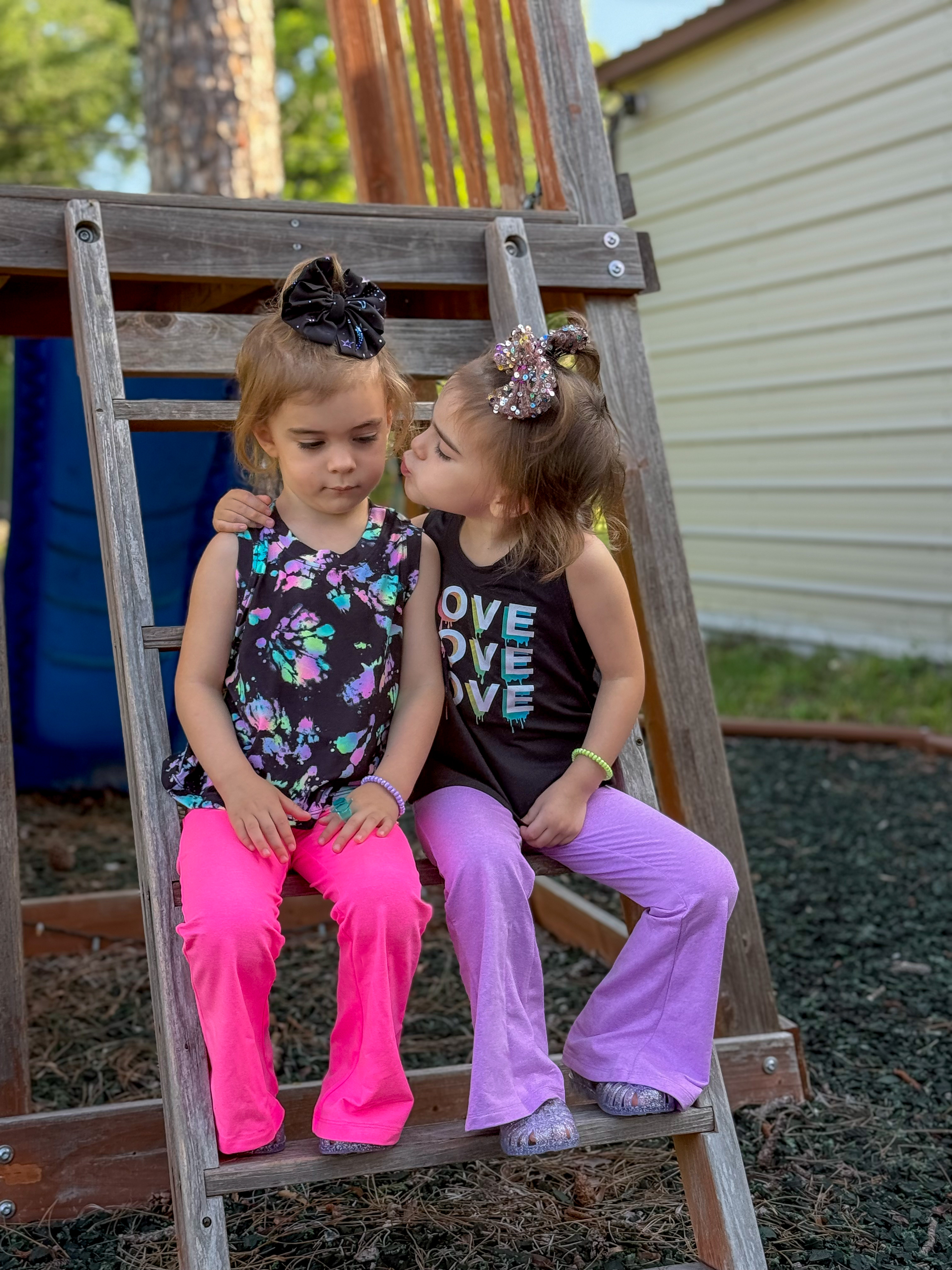 Two young children sitting on a wooden ladder, one kissing the other.