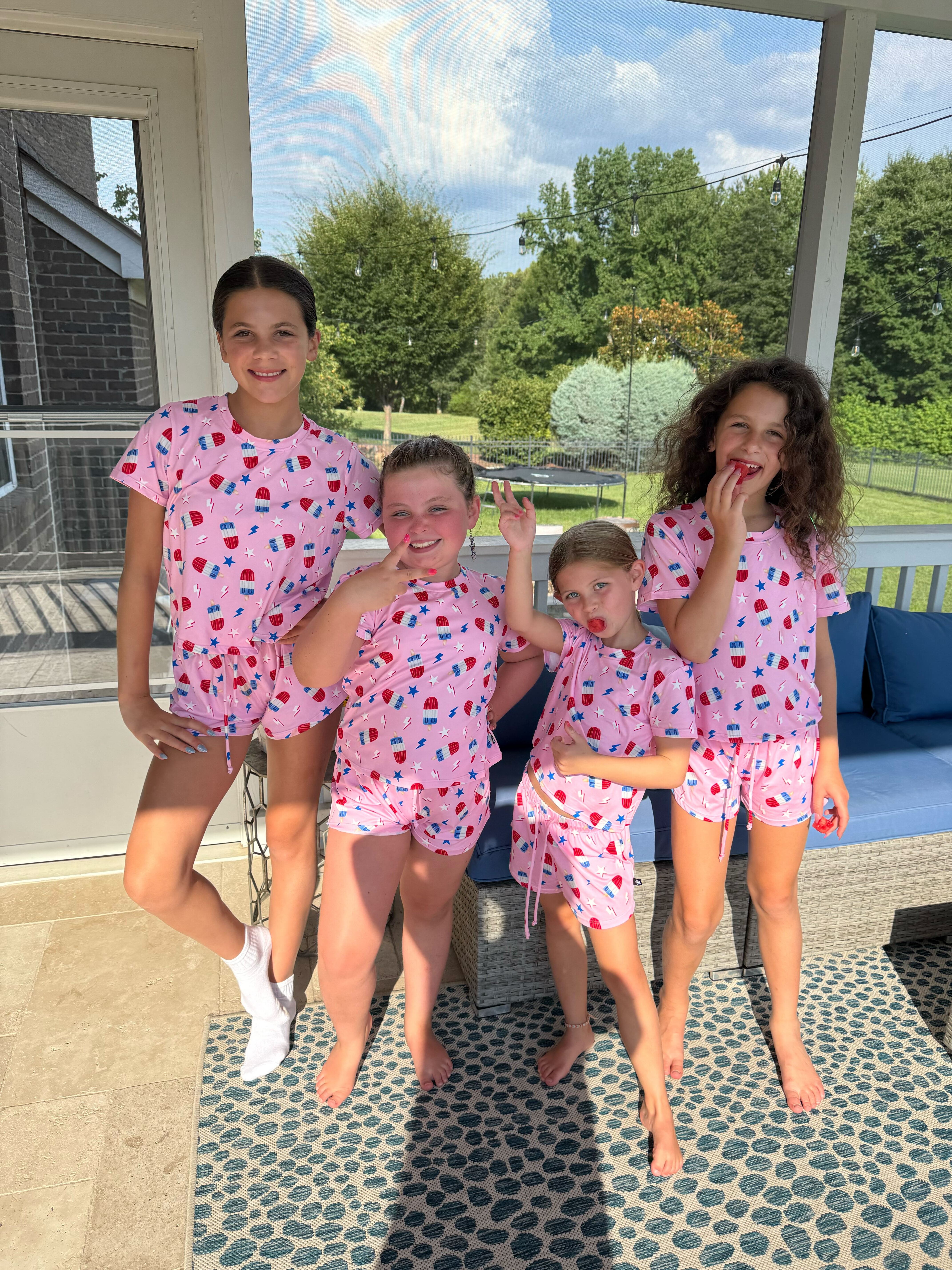 Four children in matching pink outfits on a porch, with trees in the background.