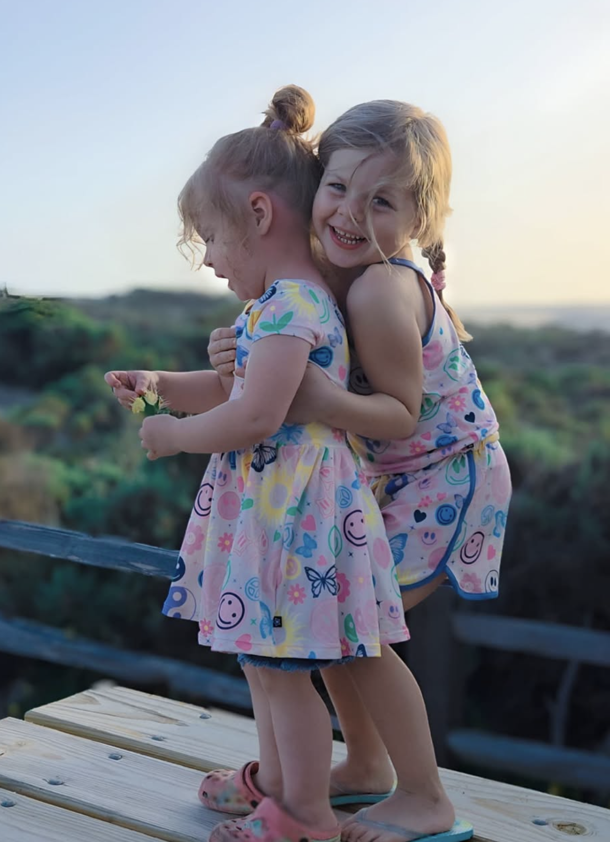 Two children in colorful outfits hug outdoors on a wooden platform.