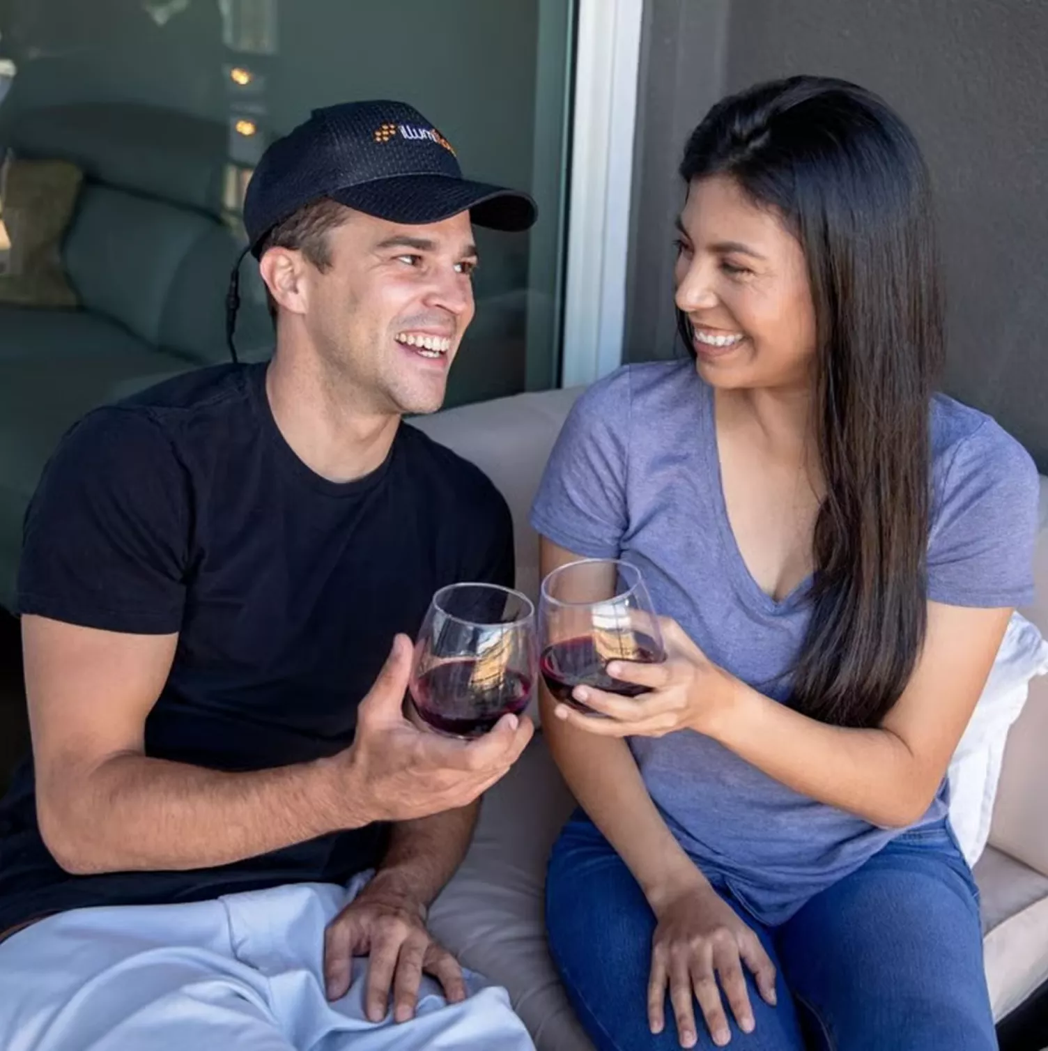 A man and a woman sit on a couch, smiling at each other while holding glasses of red wine.