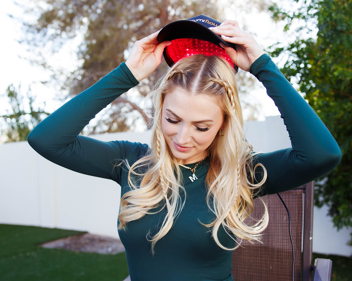 A blonde woman in a green shirt holds a black cap with red lights inside above her head.