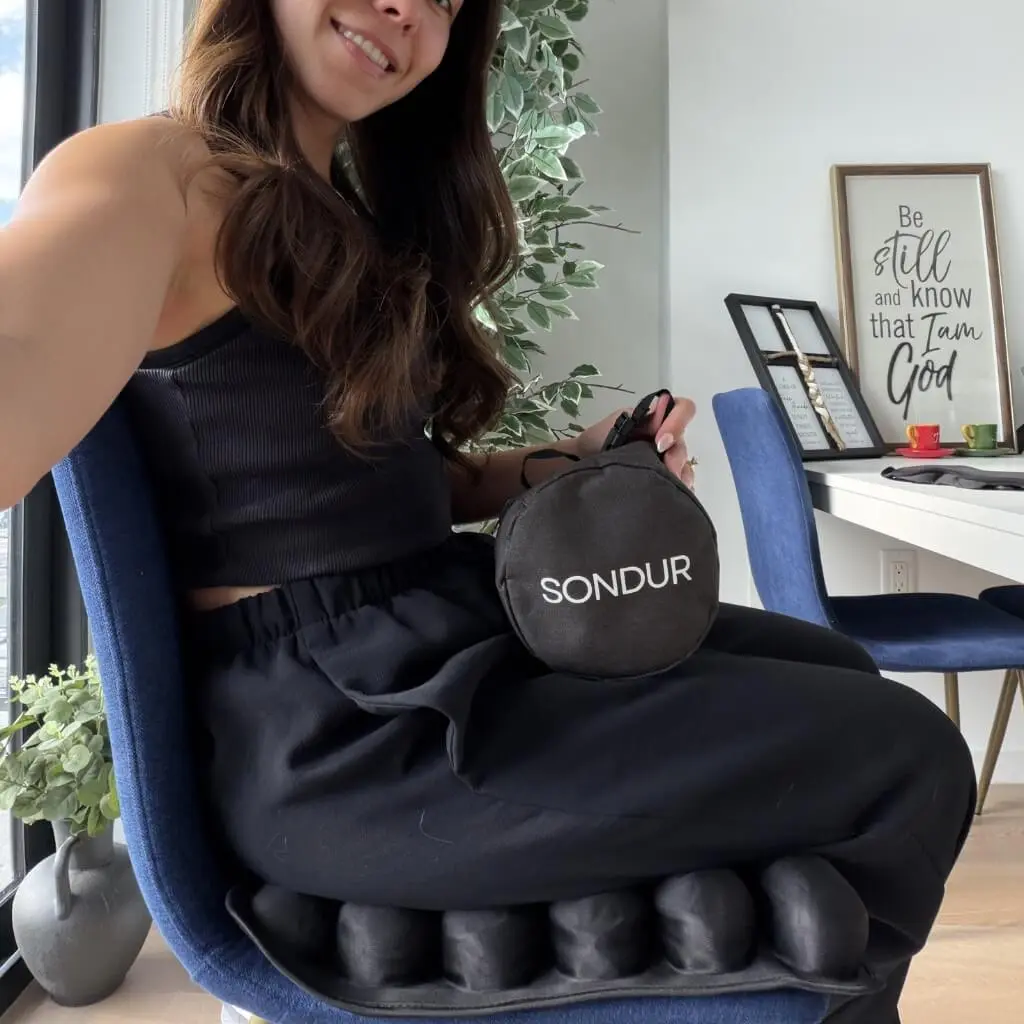 A woman sits on a black inflatable cushion on a blue chair, holding its round carrying bag.