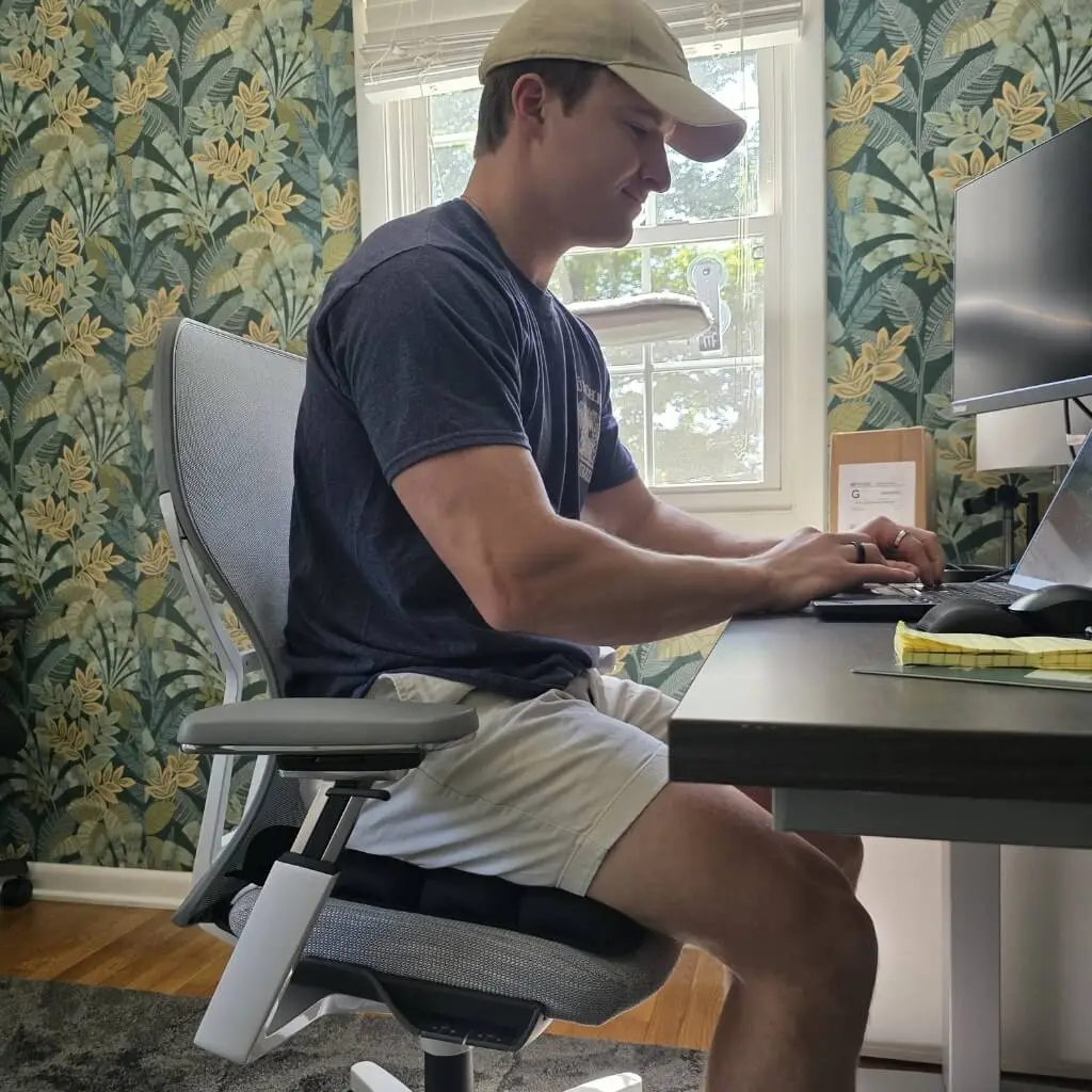 A man in a baseball cap sits on a cushion in an office chair, working on a laptop.