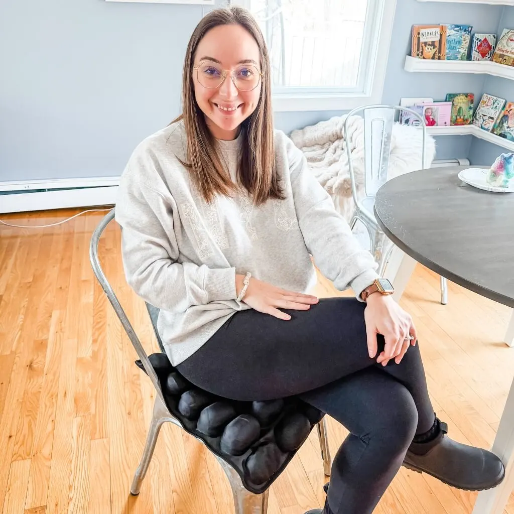 A smiling woman with glasses sits on a metal chair with a black, multi-celled seat cushion.