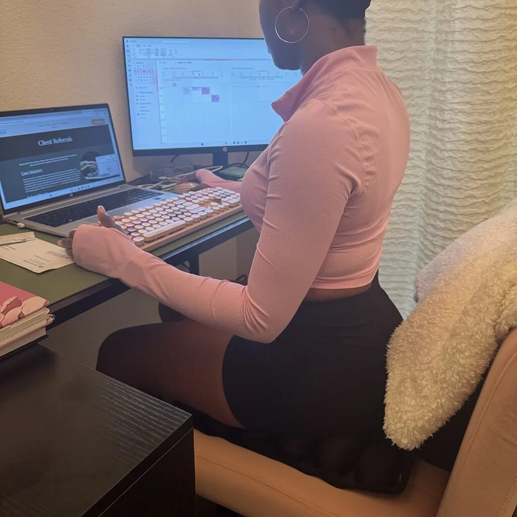 A person in a pink long-sleeved shirt sits at a desk working on a computer and laptop.