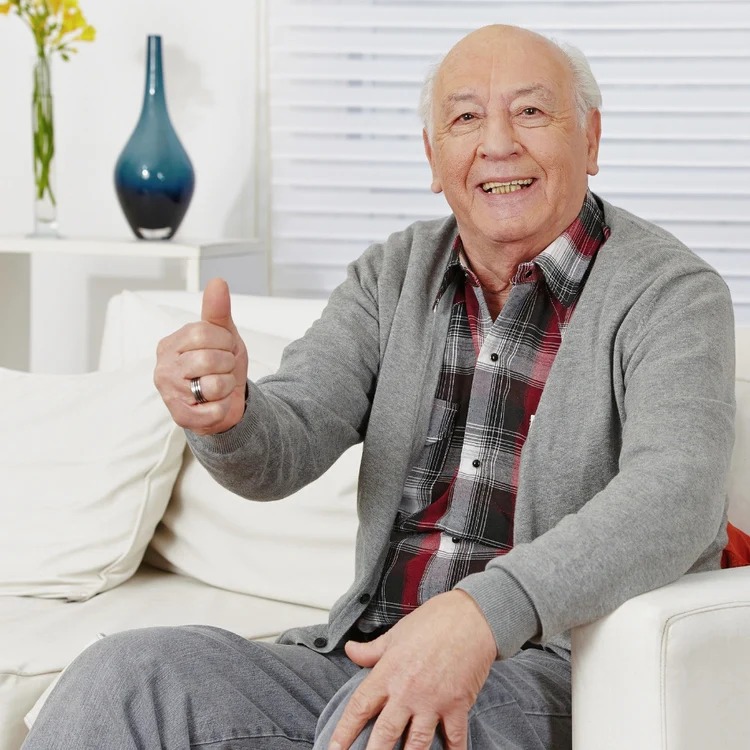 An elderly man in a grey cardigan sits on a white couch, smiling and giving a thumbs up.