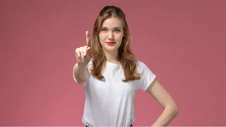 A woman in a white shirt stands against a pink background, holding up her index finger.