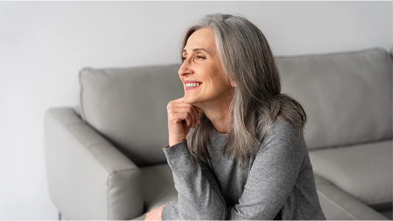 A smiling older woman with long gray hair rests her chin on her hand while sitting indoors.