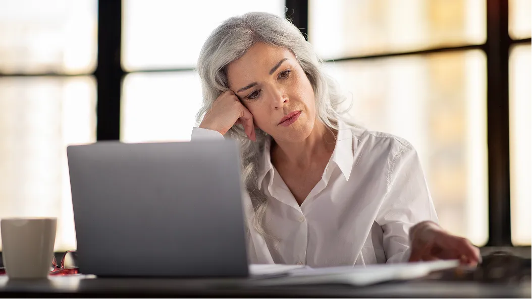 A woman with long gray hair rests her head in her hand while looking at a laptop screen.