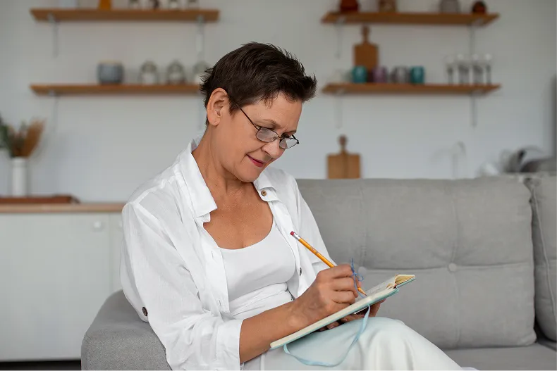 A middle-aged woman with glasses and short hair writes in a notebook while sitting on a couch.