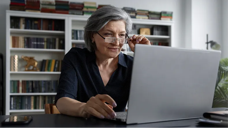 An older woman with grey hair and glasses sits at a desk, looking at a laptop computer.