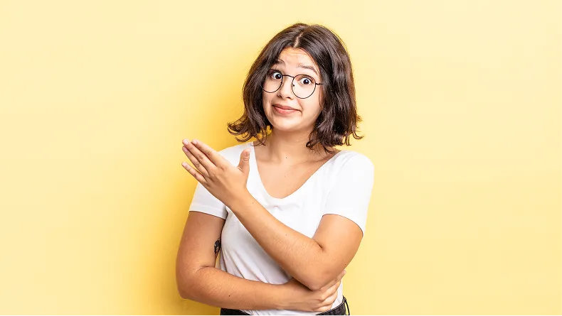 A young woman with glasses gestures to her left with a quizzical expression against a yellow background.