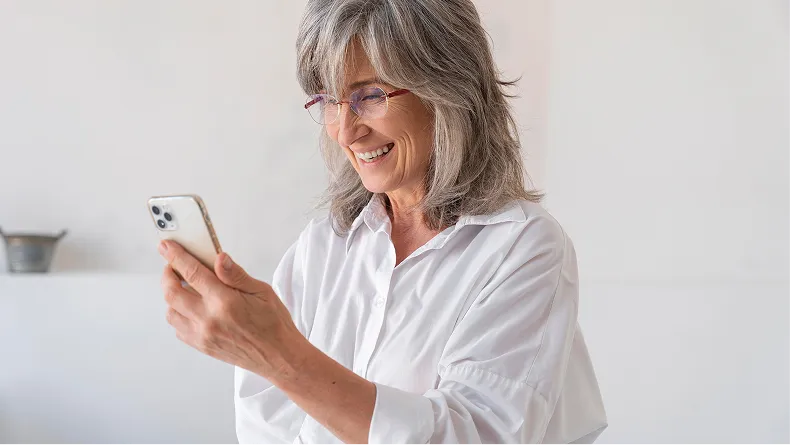 A smiling woman with grey hair and glasses, wearing a white shirt, looks at her smartphone.