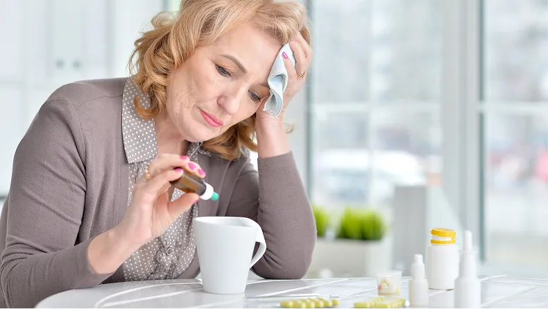 A woman who looks unwell holds a cloth to her forehead while adding drops to a mug.