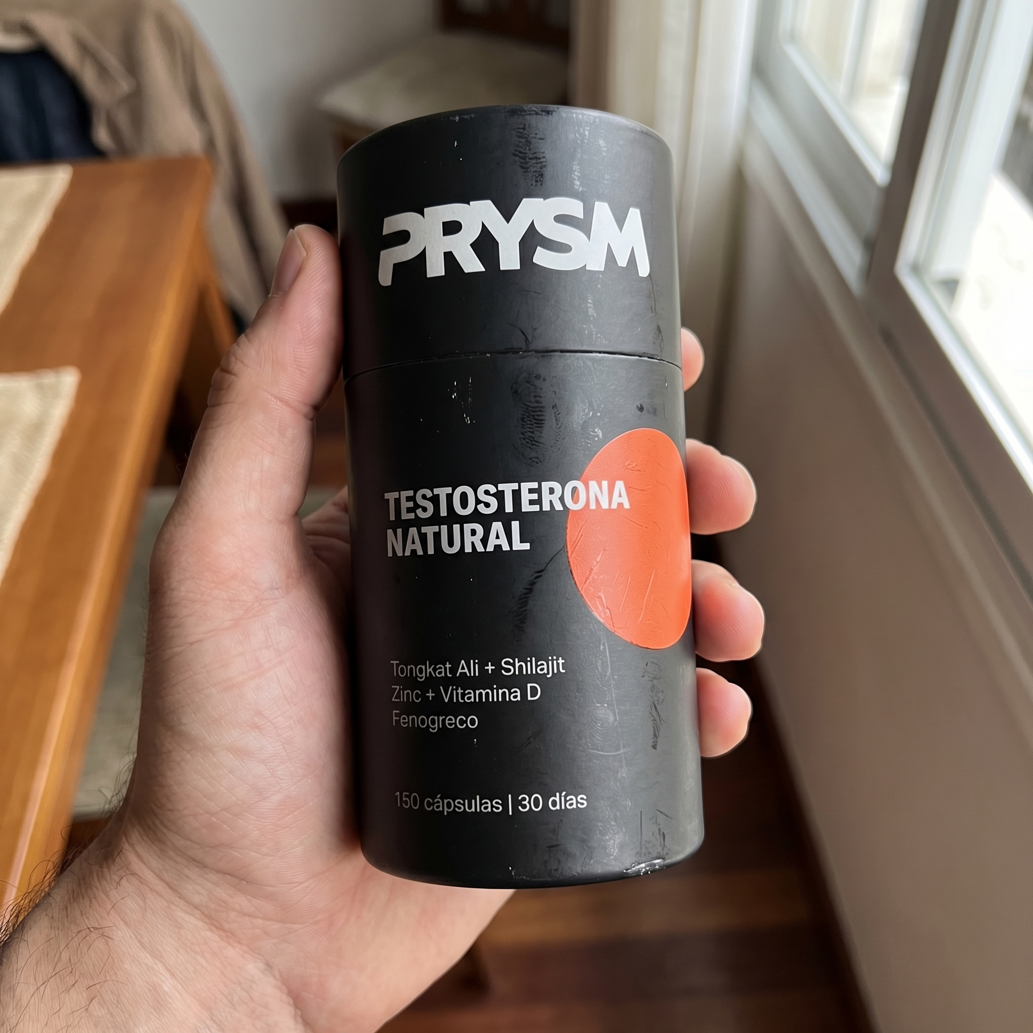 A person's hand holding a black cylindrical container of Prysm brand 'Testosterona Natural' supplements.