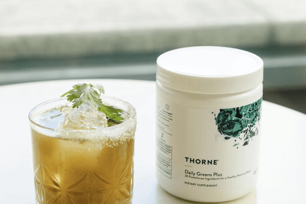 A white jar of Thorne Daily Greens Plus sits next to a green beverage in a garnished, sugar-rimmed glass.