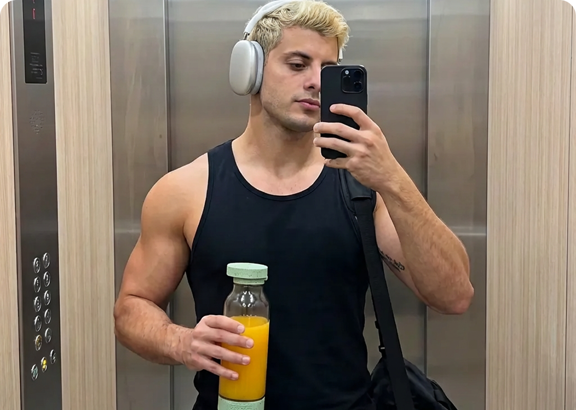 Person in an elevator taking a mirror selfie holding juice and wearing headphones.