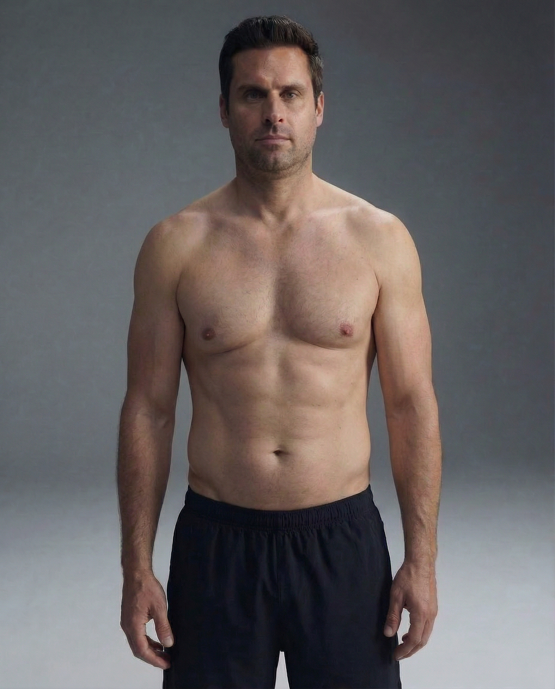 A shirtless man in black athletic shorts stands facing forward against a solid gray background.