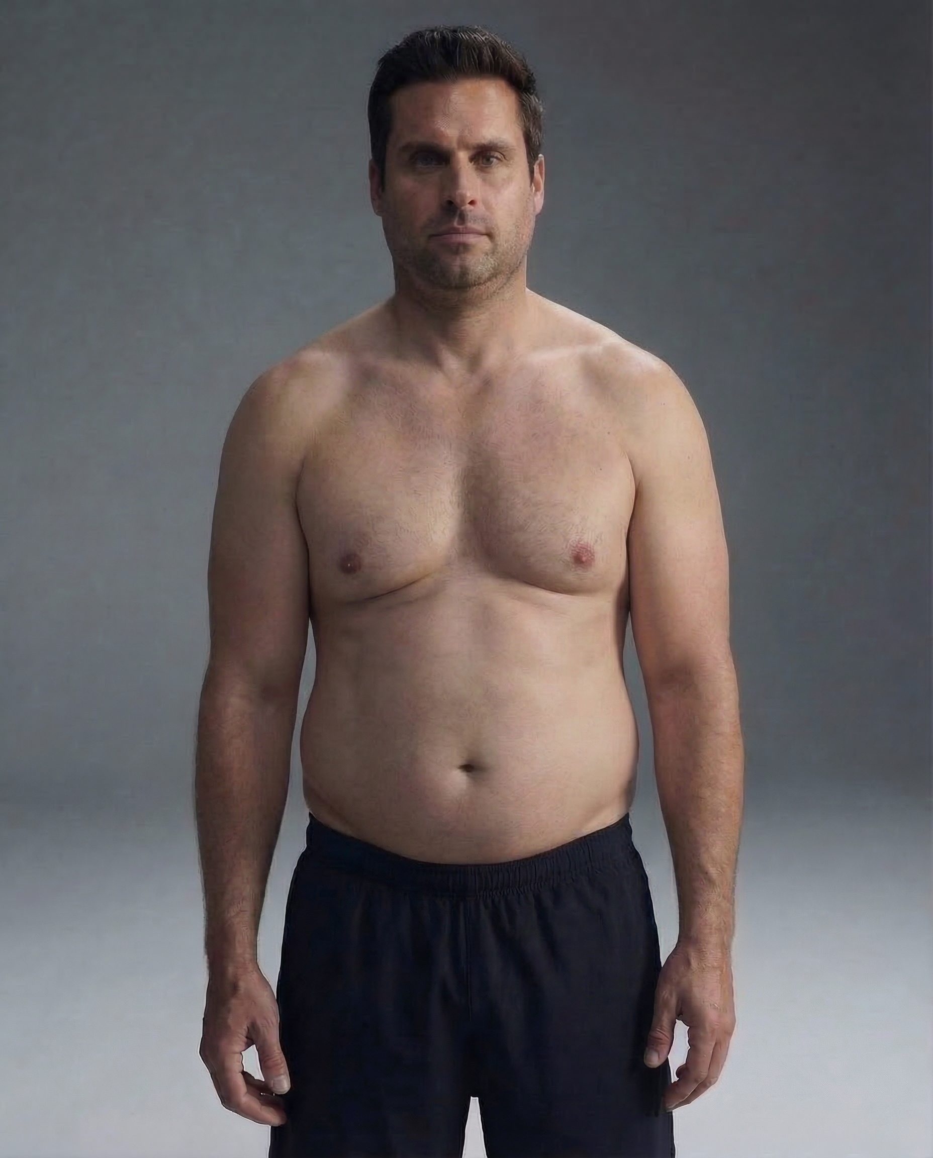 A shirtless man in black shorts stands in front of a plain grey background, facing forward.