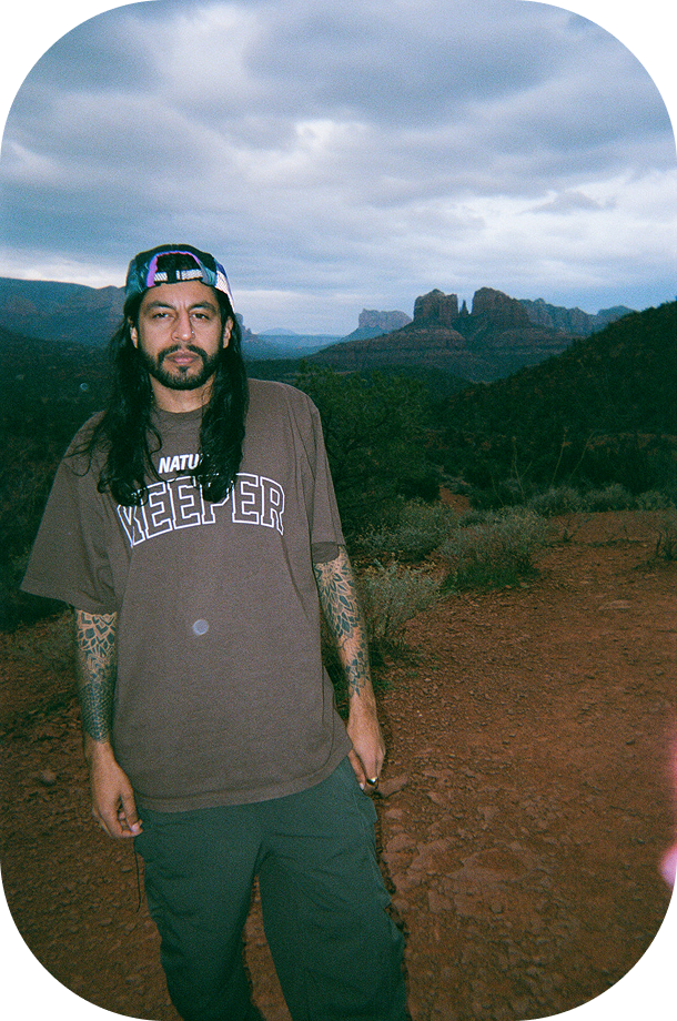 A person with long hair and tattooed arms stands on a trail overlooking desert mountains at dusk.