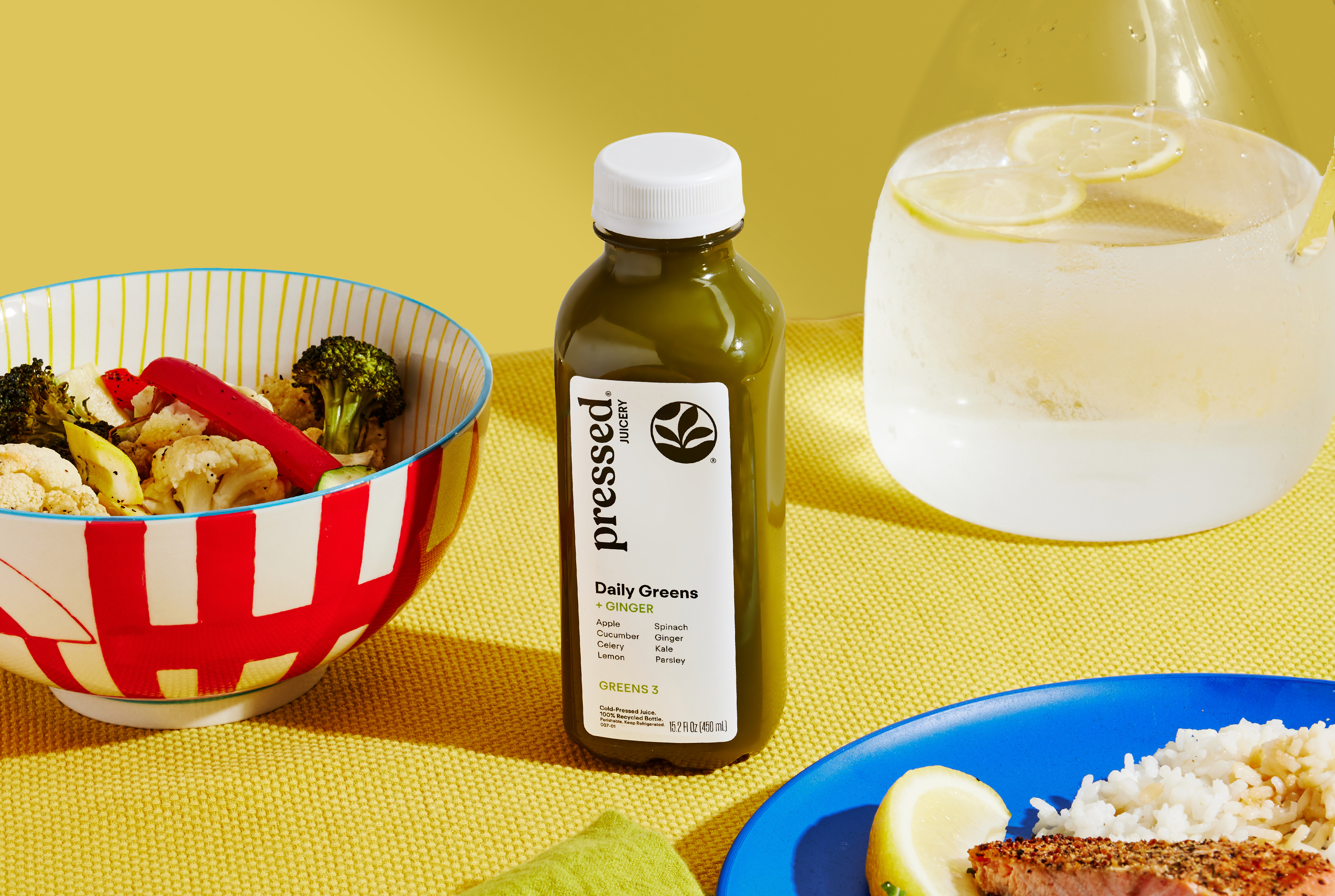 A bottle of green juice sits on a yellow placemat next to a bowl of vegetables and a plate of salmon.