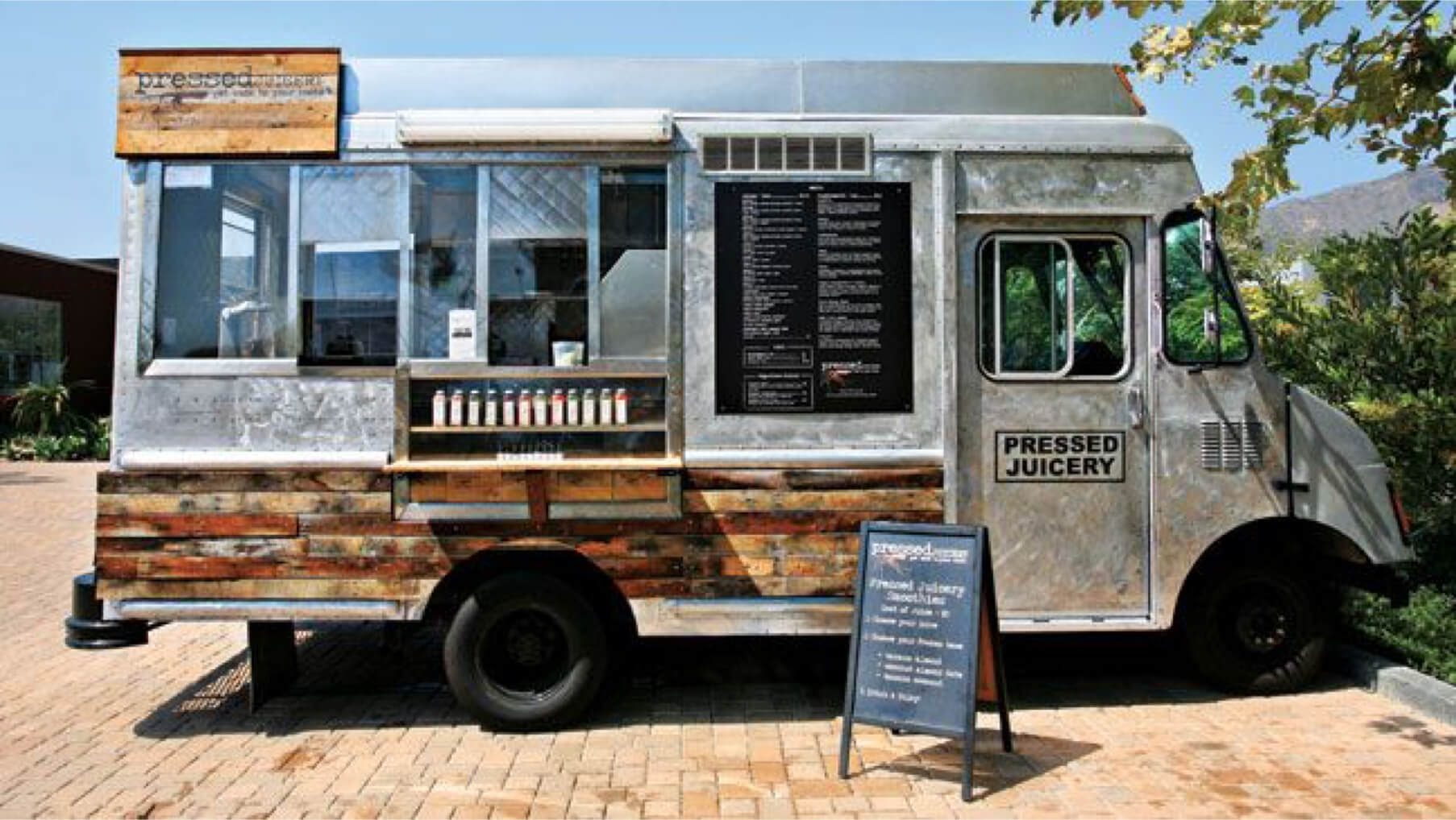 A silver and wood-paneled 'Pressed Juicery' food truck parked outdoors on a paved lot.