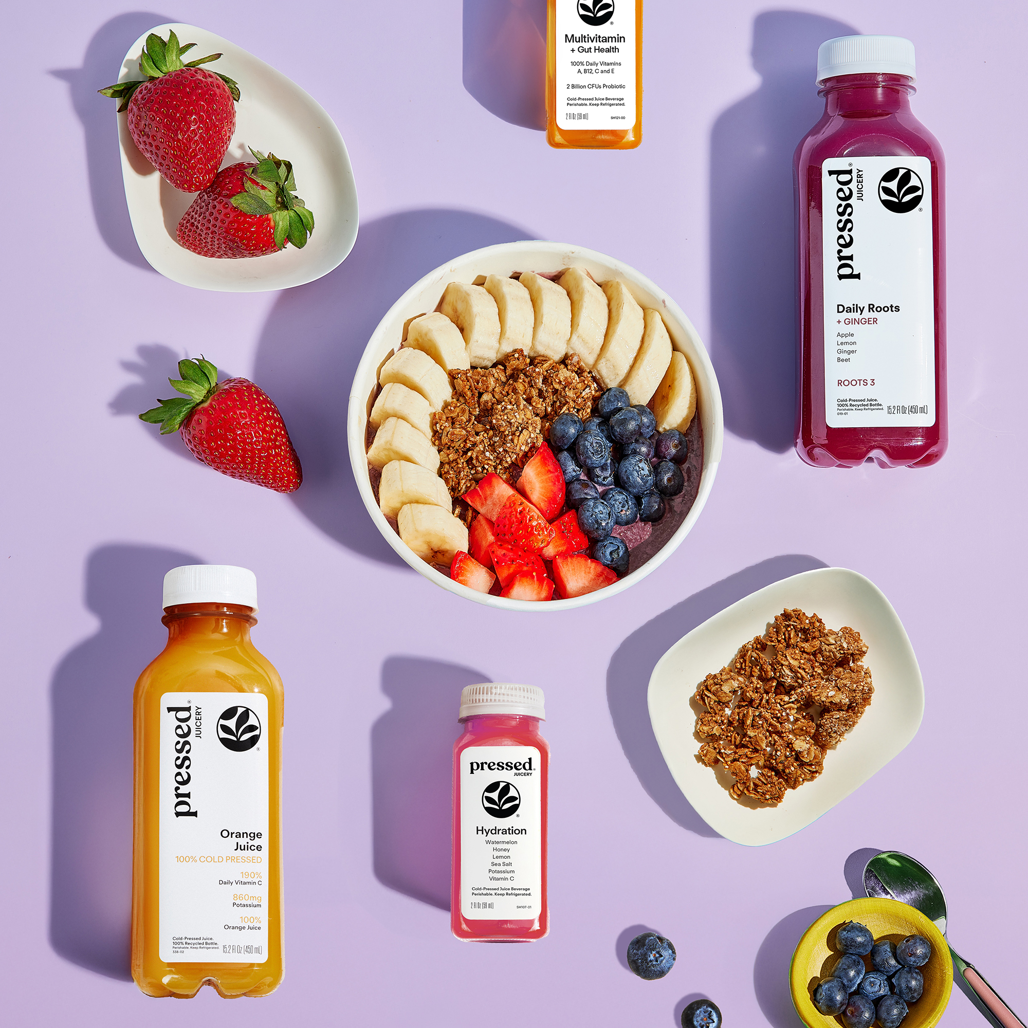 An overhead shot of Pressed juices and a fruit and granola bowl on a purple background.