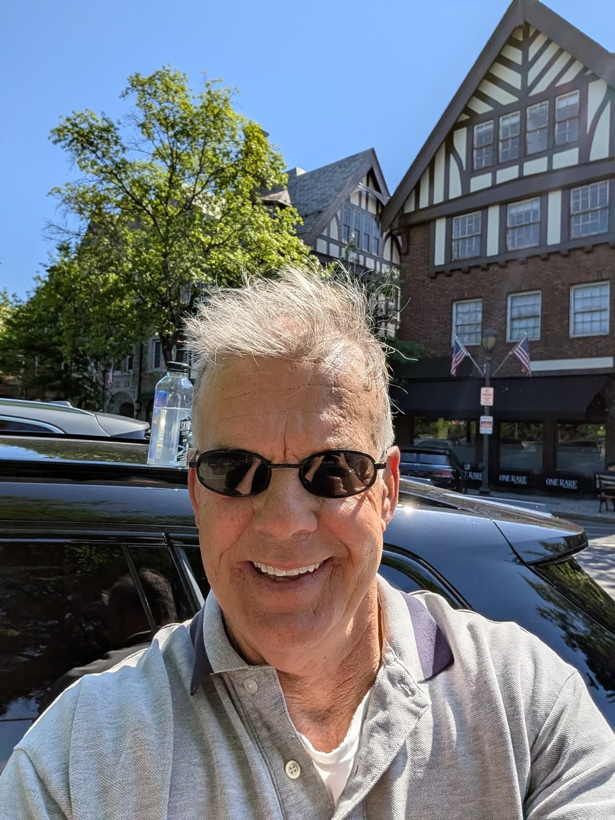 A smiling man with gray hair and sunglasses takes a selfie outdoors on a sunny day.