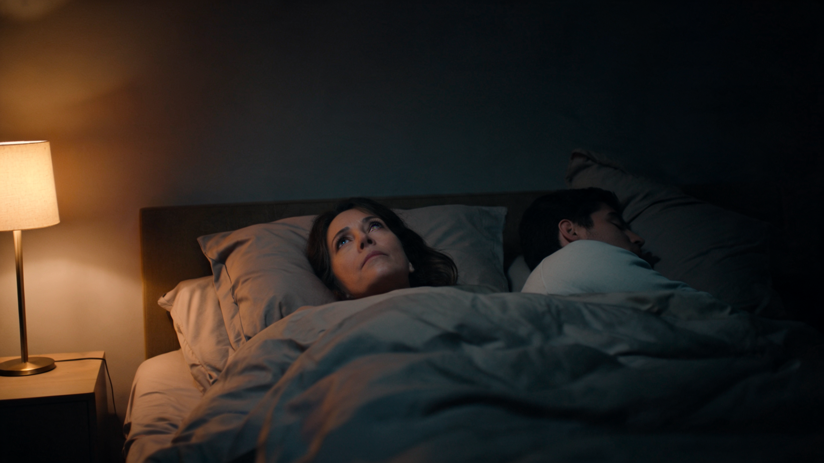 Woman lying awake in bed at night while her partner sleeps peacefully beside her, illuminated by a warm bedside lamp.